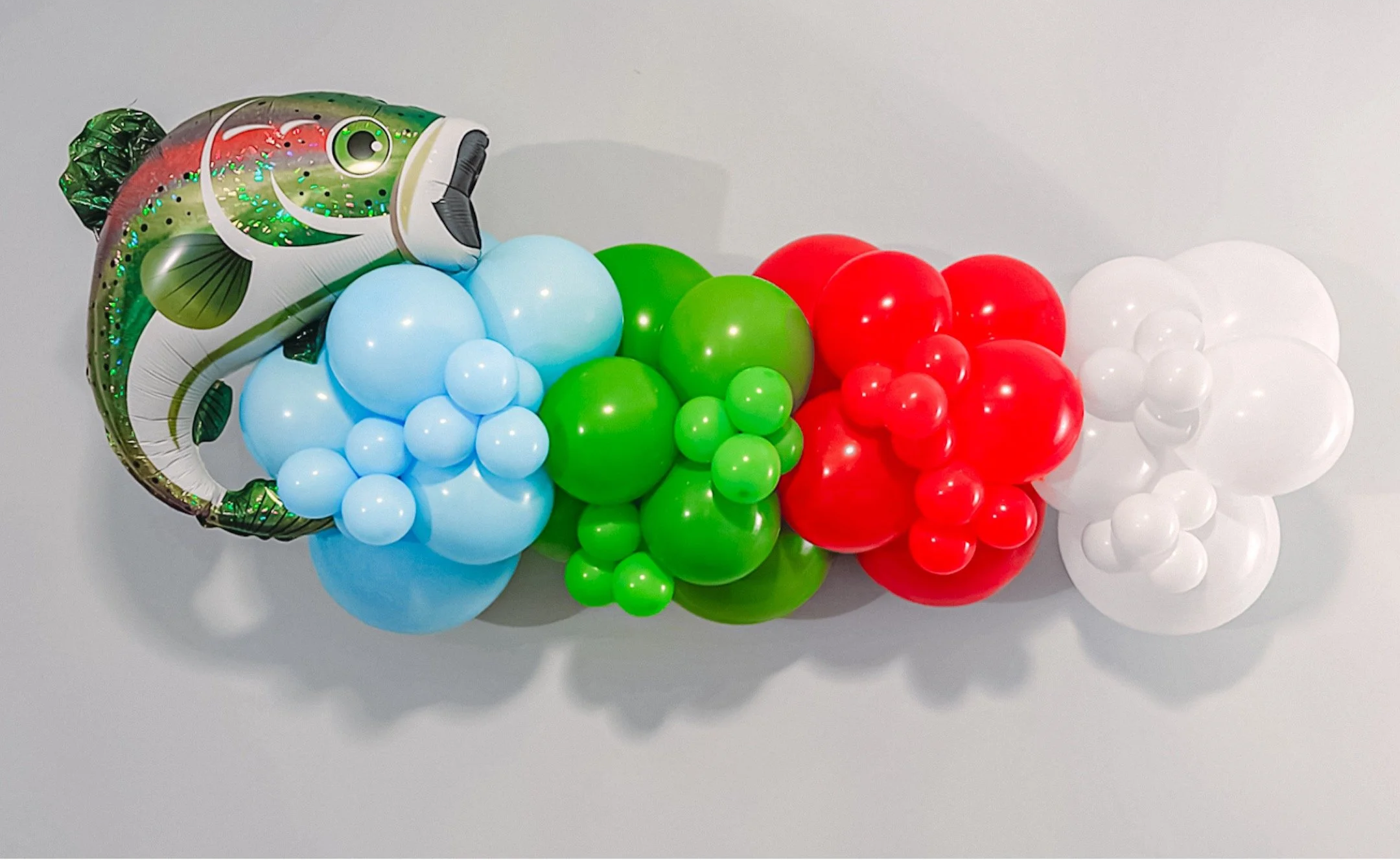 Colorful balloon arrangement with a fish-shaped foil balloon at the left, featuring blue, green, red, and white balloons grouped in clusters.
