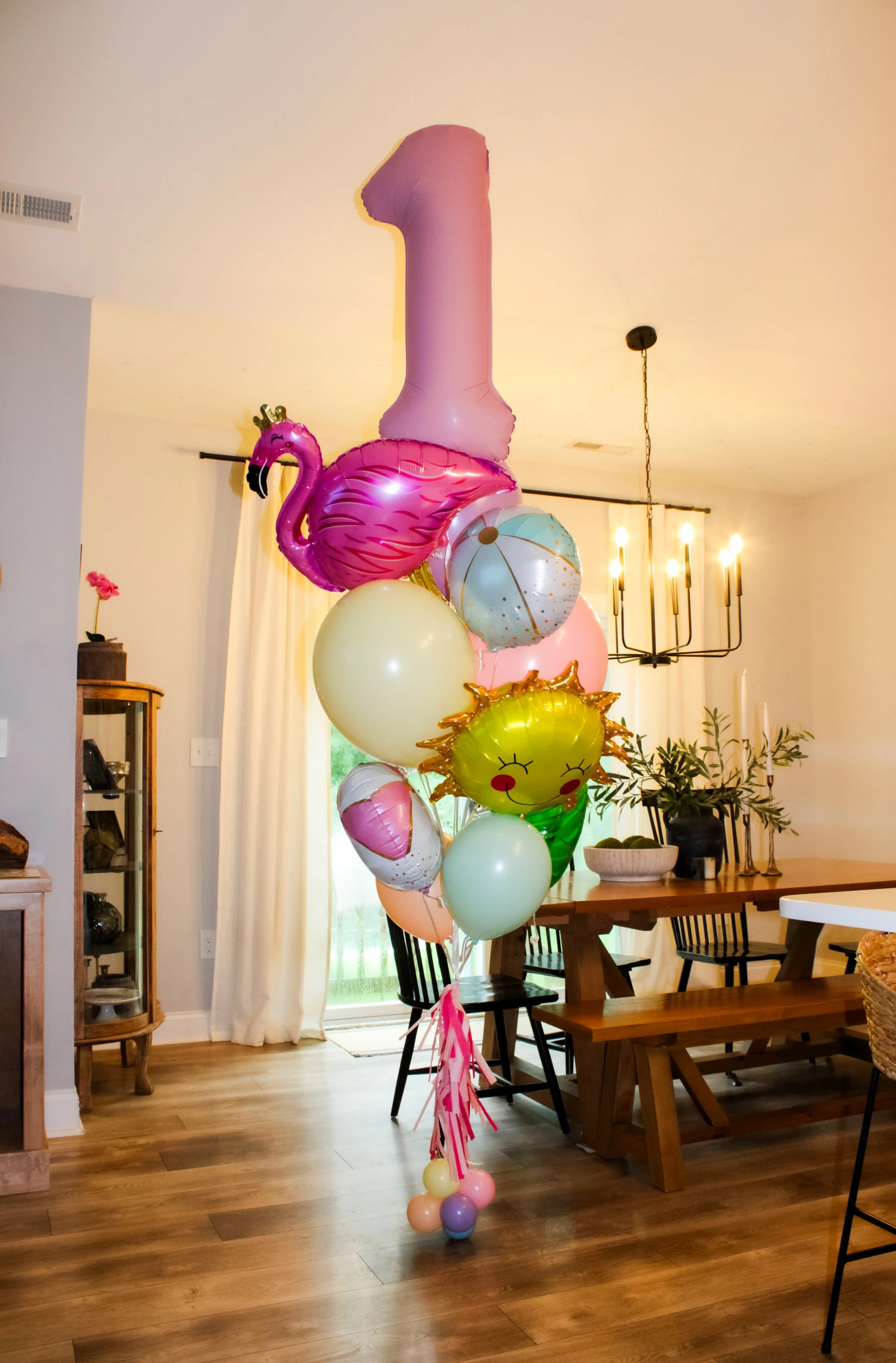 A cluster of colorful balloons for a birthday celebration includes a pink number one balloon, a pink flamingo with a crown, a sun with a smiling face, a beach ball, and various pastel-colored balloons, all tied together and placed in a spacious livin