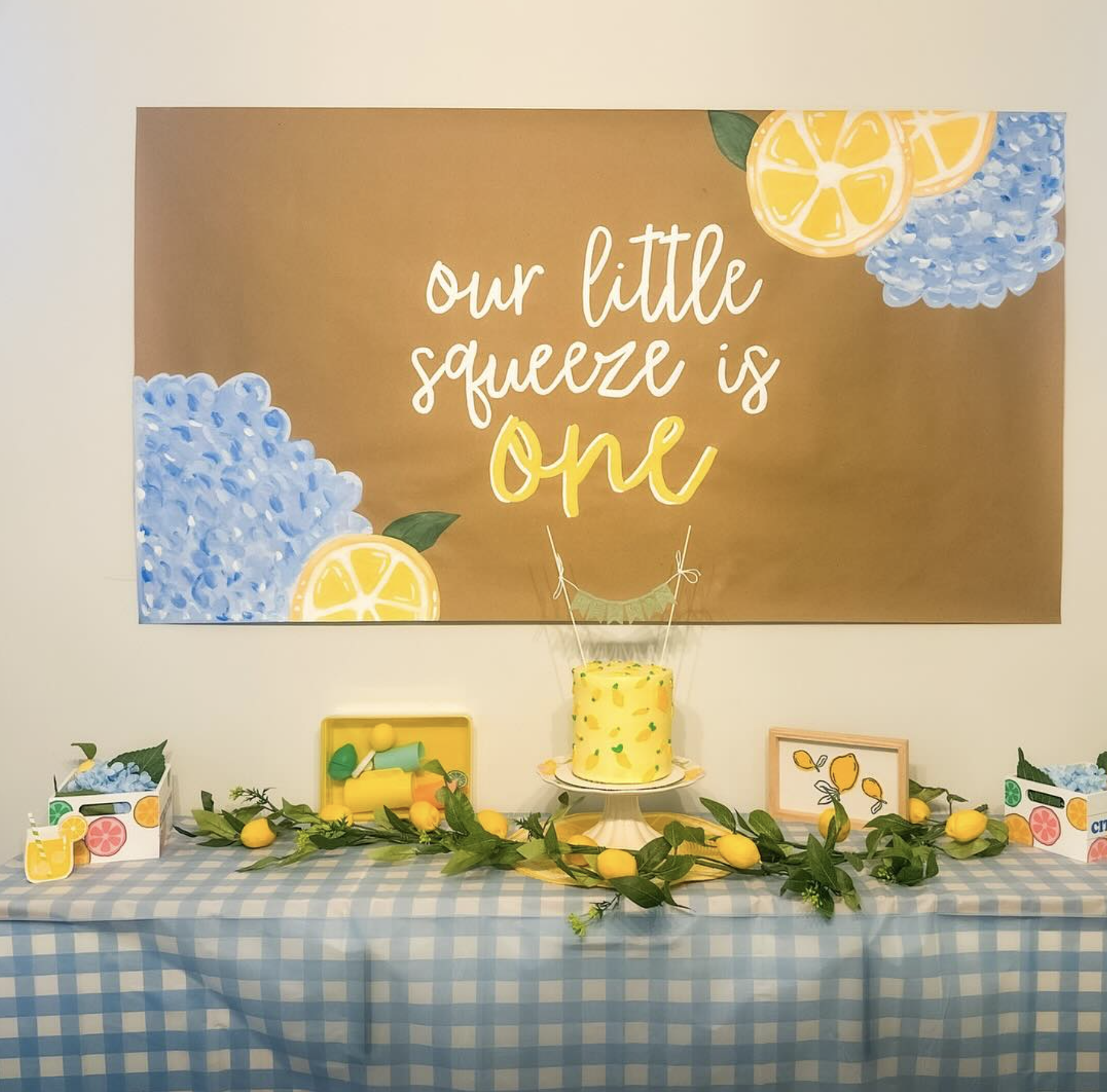 Decorated table with a yellow cake, decorations, and lemon-themed items, with a large poster behind that reads 'our little squeeze is one'.