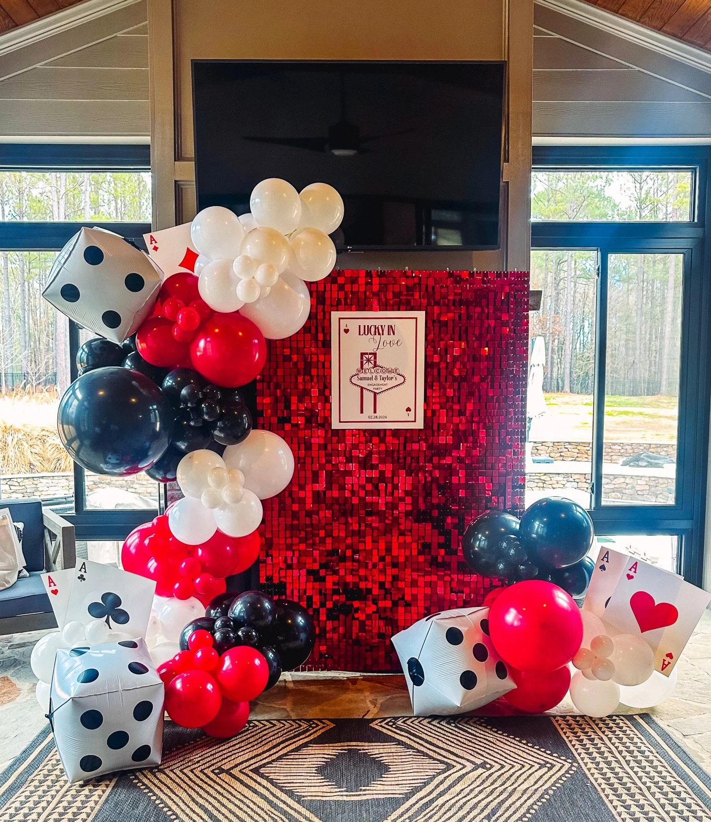 Valentine's Day-themed photo backdrop with red and black balloons, giant playing cards, and a red sequin backdrop, with a sign in the center that reads 'Lucky in Love' and includes a personalized message and date.