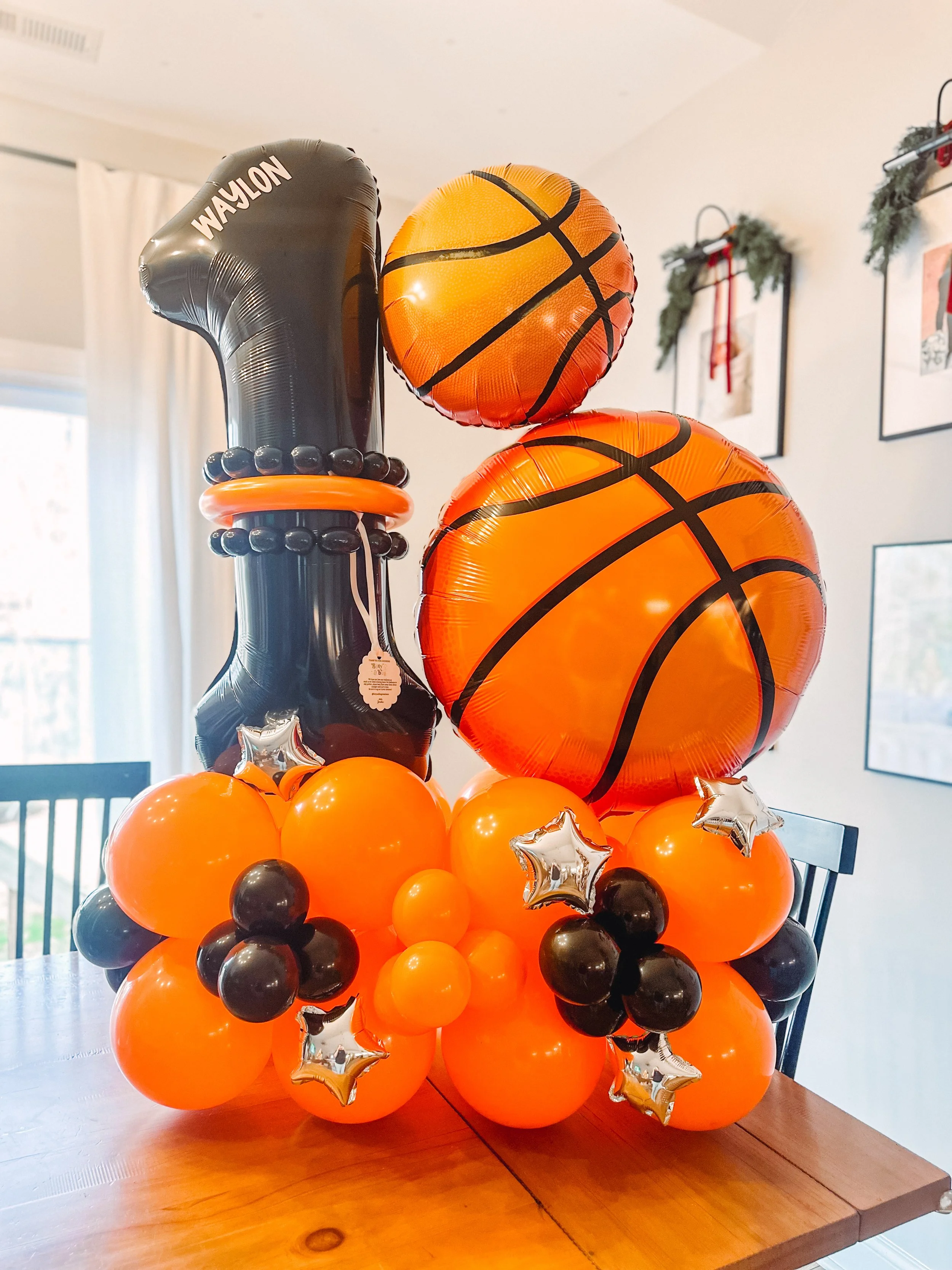 Basketball-themed balloon arrangement with orange and black basketball balloons, star-shaped silver balloons, and a large black balloon that says 'WAYLON'