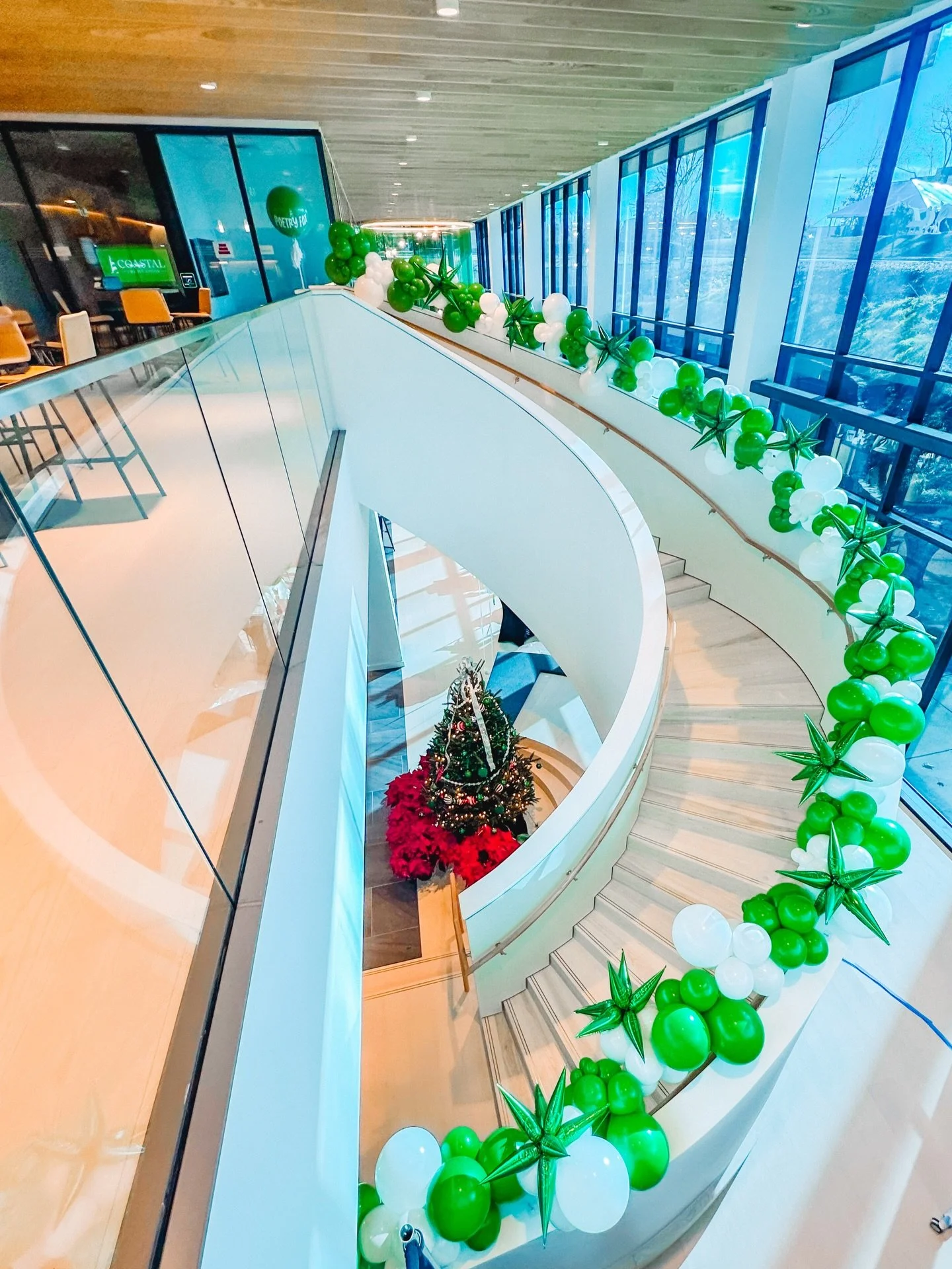 A modern indoor space with a spiral staircase decorated with green and white balloons, a Christmas tree at the bottom, and large floor-to-ceiling windows letting in natural light.