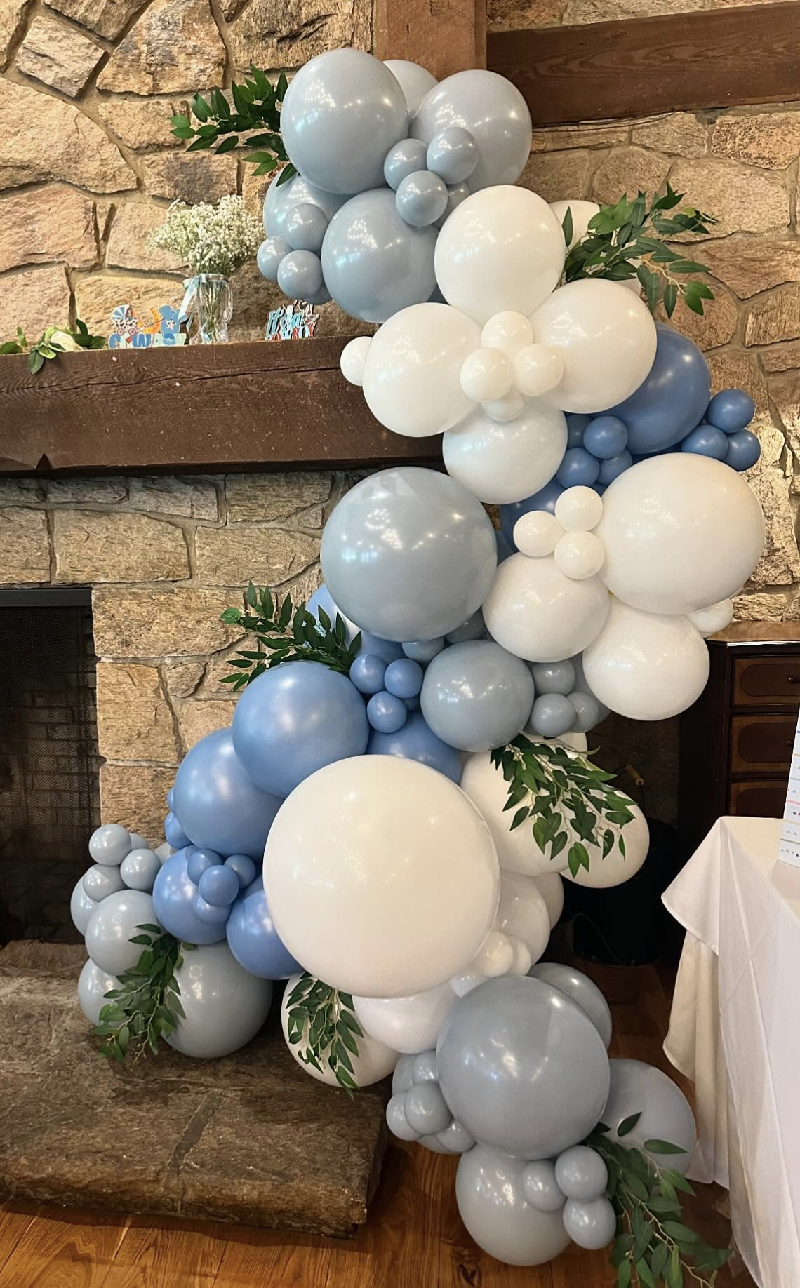 Decorative balloon arrangement with white and light blue balloons and green leaves, positioned beside a stone fireplace in a room with wooden floors.