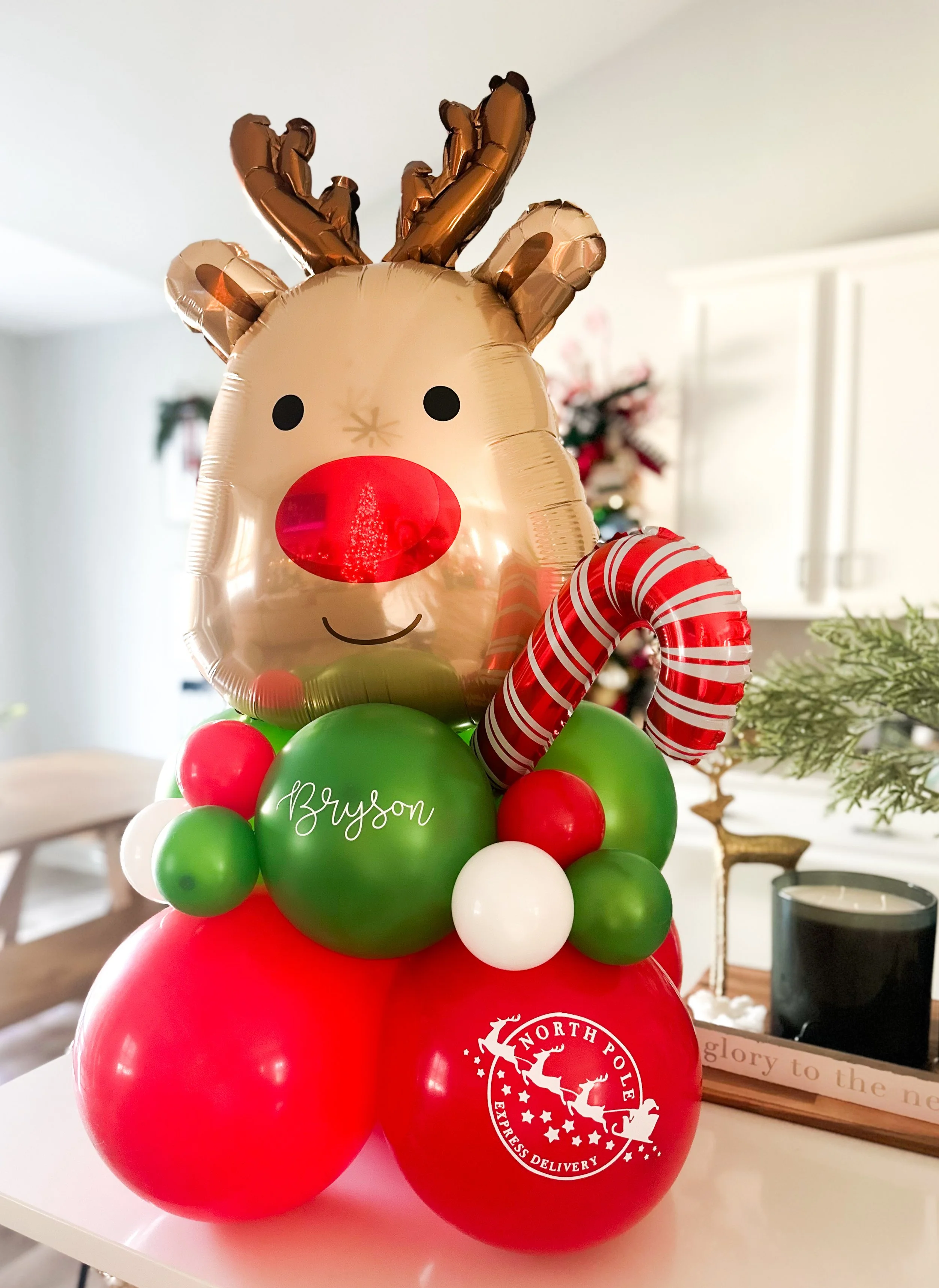 Christmas-themed balloon arrangement with a reindeer face balloon, red, green, and white balloons, a candy cane shaped balloon, and a balloon with the name "Bryson" on it.