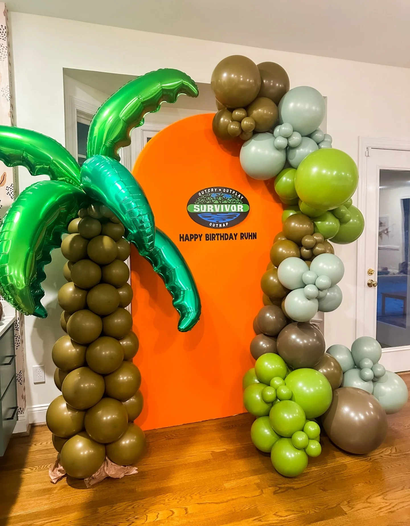 Balloon arch with green palm trees and a Survivor-themed backdrop for a birthday celebration.