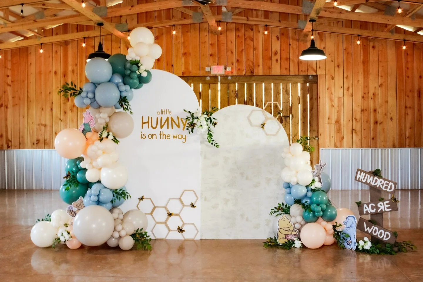Birthday party decoration with a honey theme, featuring a balloon arch, honey bee and Winnie the Pooh cutouts, and a sign reading 'Hundy, Ace, Wood' in a wooden barn-like venue.