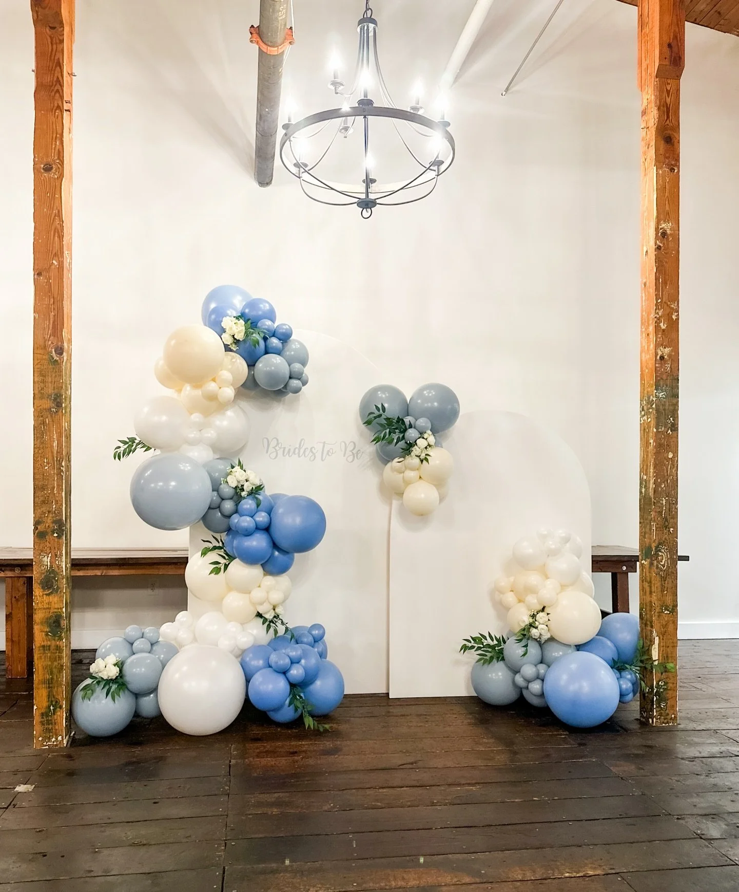 Balloon decoration with clusters of white, blue, and gray balloons, with some greenery and white flowers, set against a white backdrop with a chandelier overhead, for a bridal or baby shower celebration.