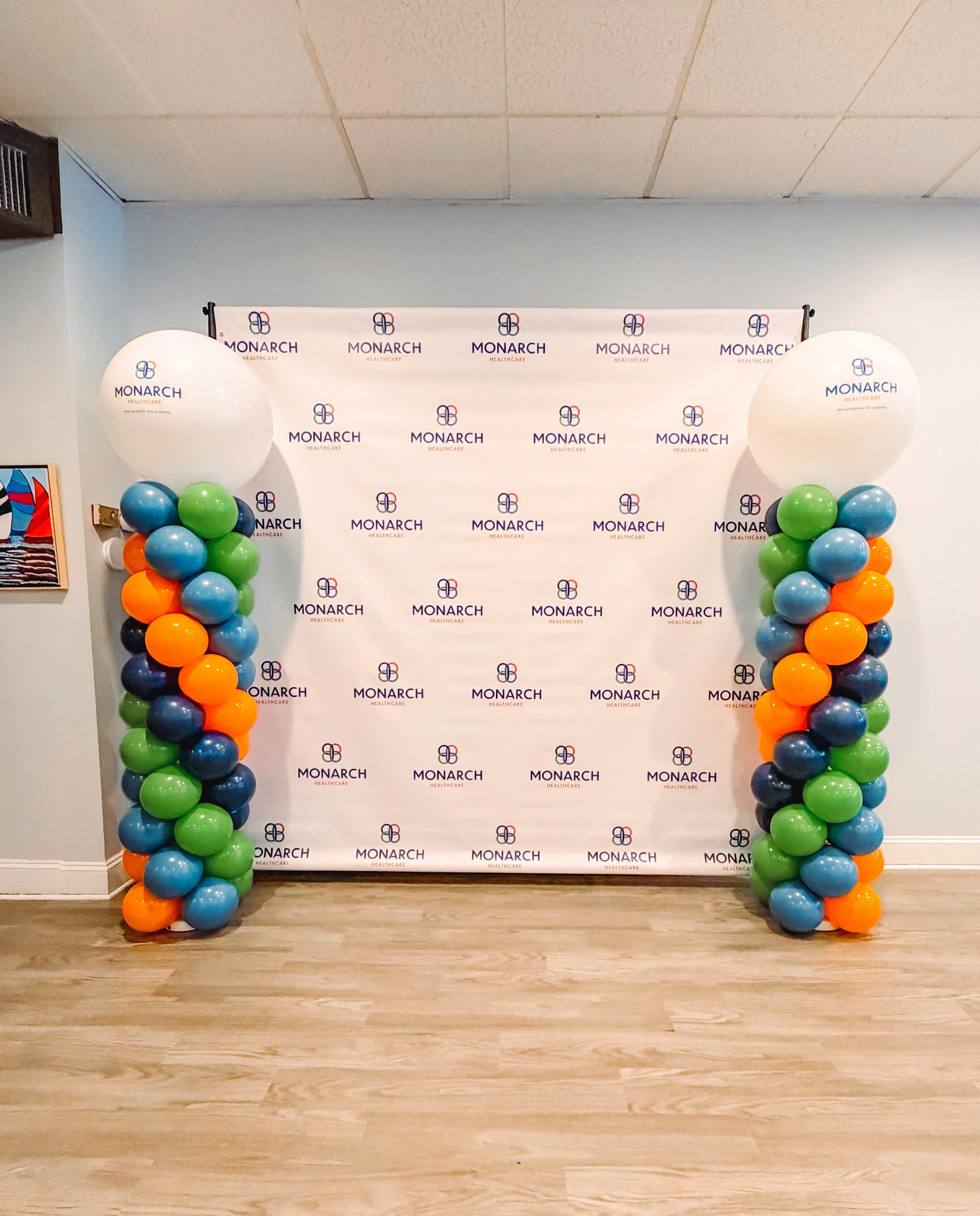 A backdrop with the Monarch Healthcare logo and text, flanked by two columns of balloons in orange, blue, green, gray, and black, on a wooden floor and a plain wall.