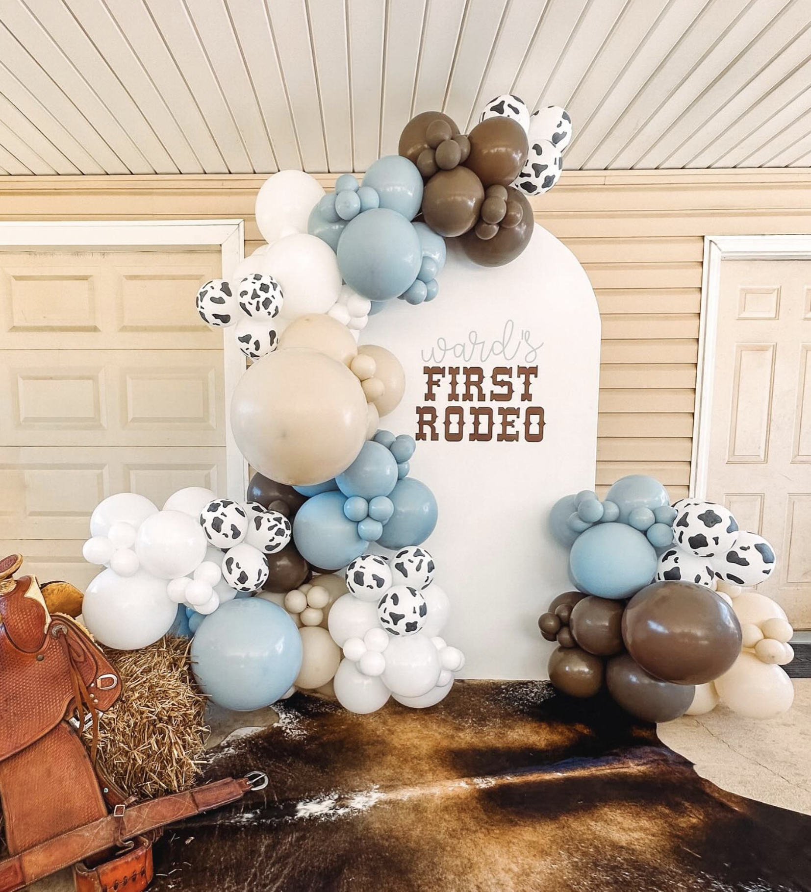 Decorative balloon arrangement with a western theme for a child's first rodeo celebration, featuring various shades of blue, white, brown, and cow print balloons, with a white backdrop that reads "Ward's First Rodeo."