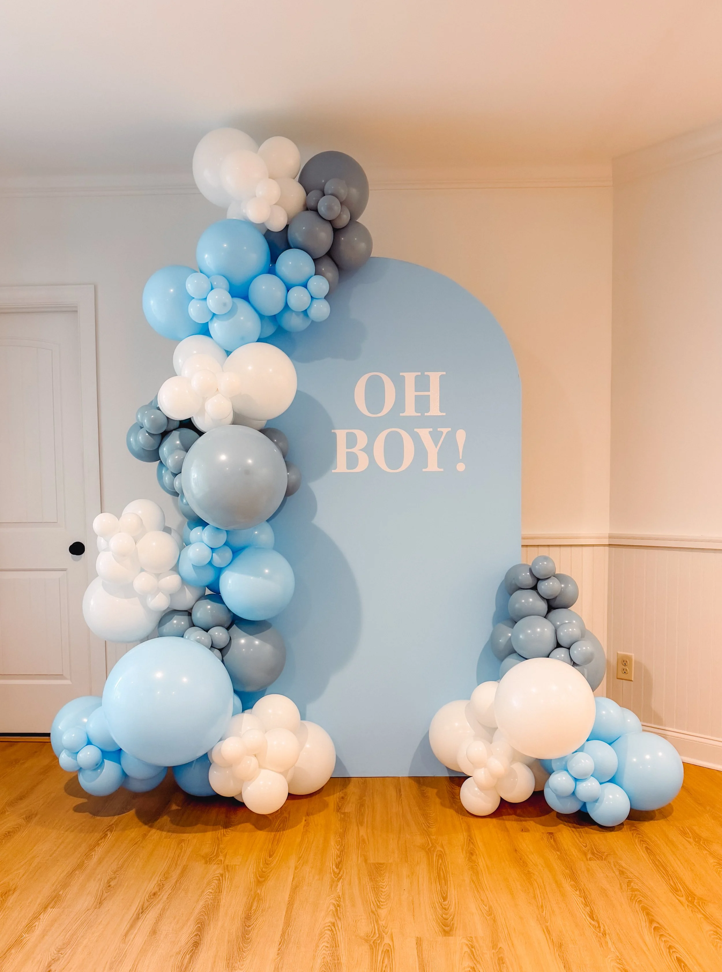 Balloon arrangement with white, blue, gray, and black balloons surrounding a blue backdrop that reads 'OH BOY!' for a baby shower or gender reveal celebration.