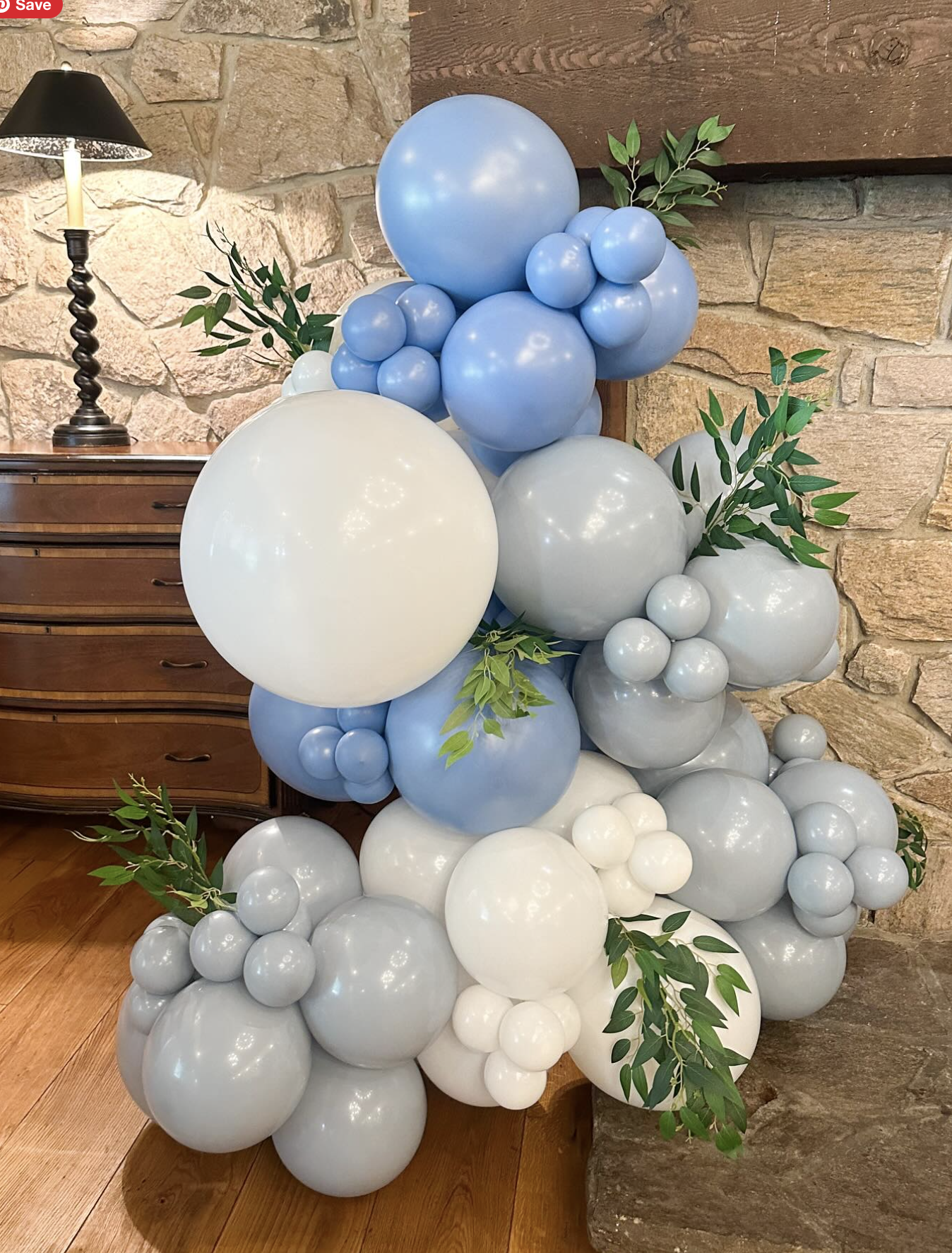 Arrangement of blue, white, and light gray balloons with green leafy accents near a stone wall and wooden furniture.