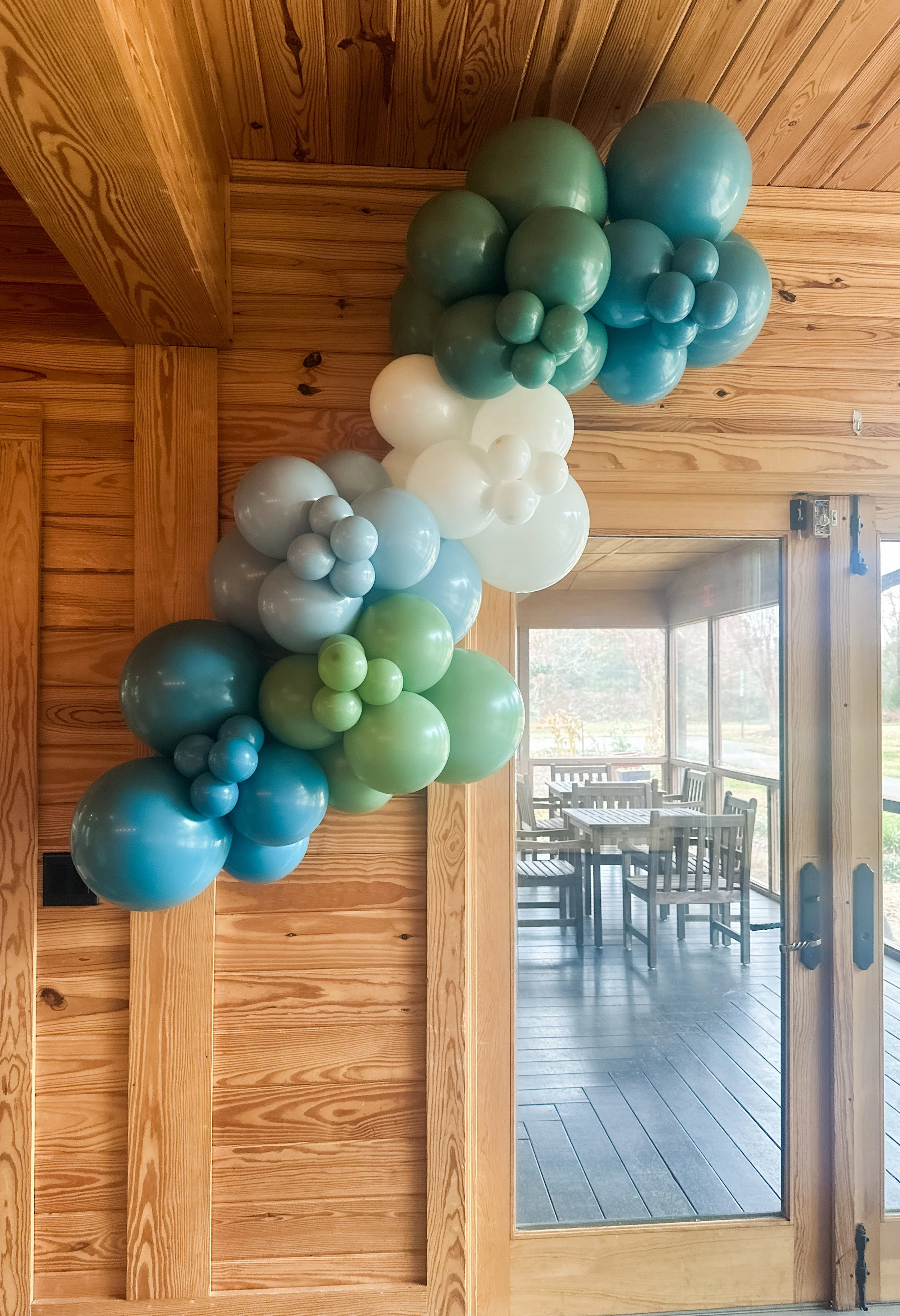 A balloon garland with shades of blue, green, white, and gray, arranged on a wooden wall near a sliding glass door leading to an outdoor patio.