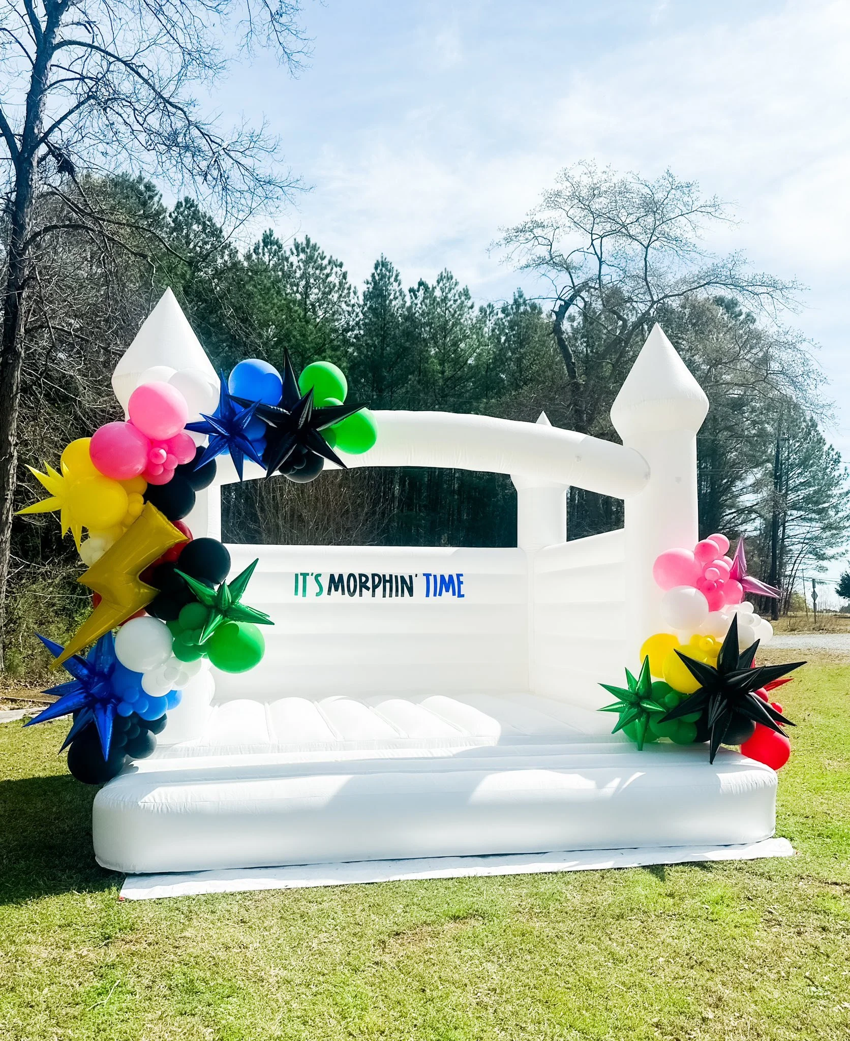Inflatable white castle with colorful balloons and the phrase 'It's Morphin' Time' displayed on it, set outdoors on grass with trees and blue sky in the background.