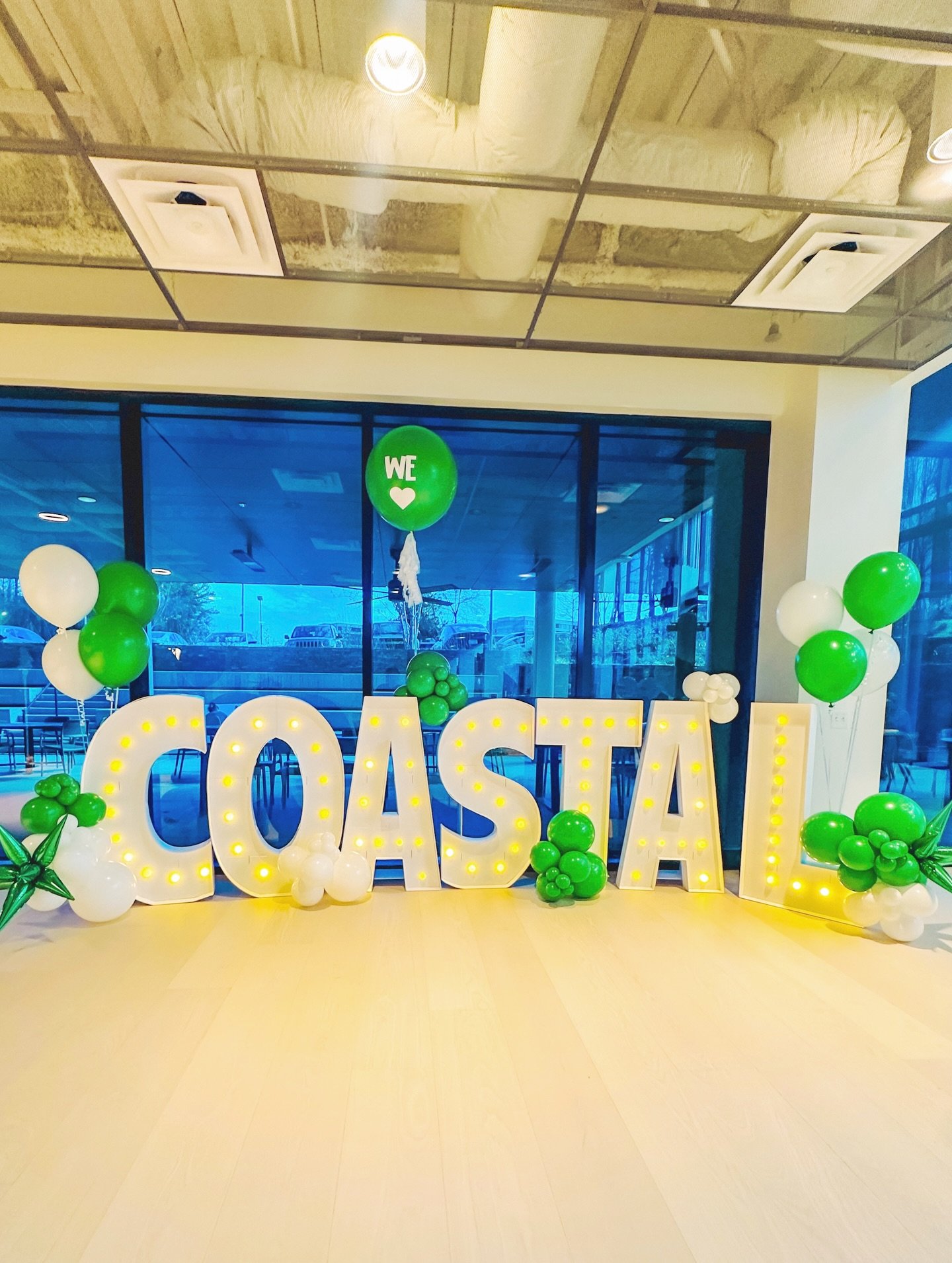 Decorative display with large illuminated white letters spelling 'COASTAL' surrounded by green and white balloons and a green balloon with 'WE ♥' on it, inside an indoor space with large glass windows.