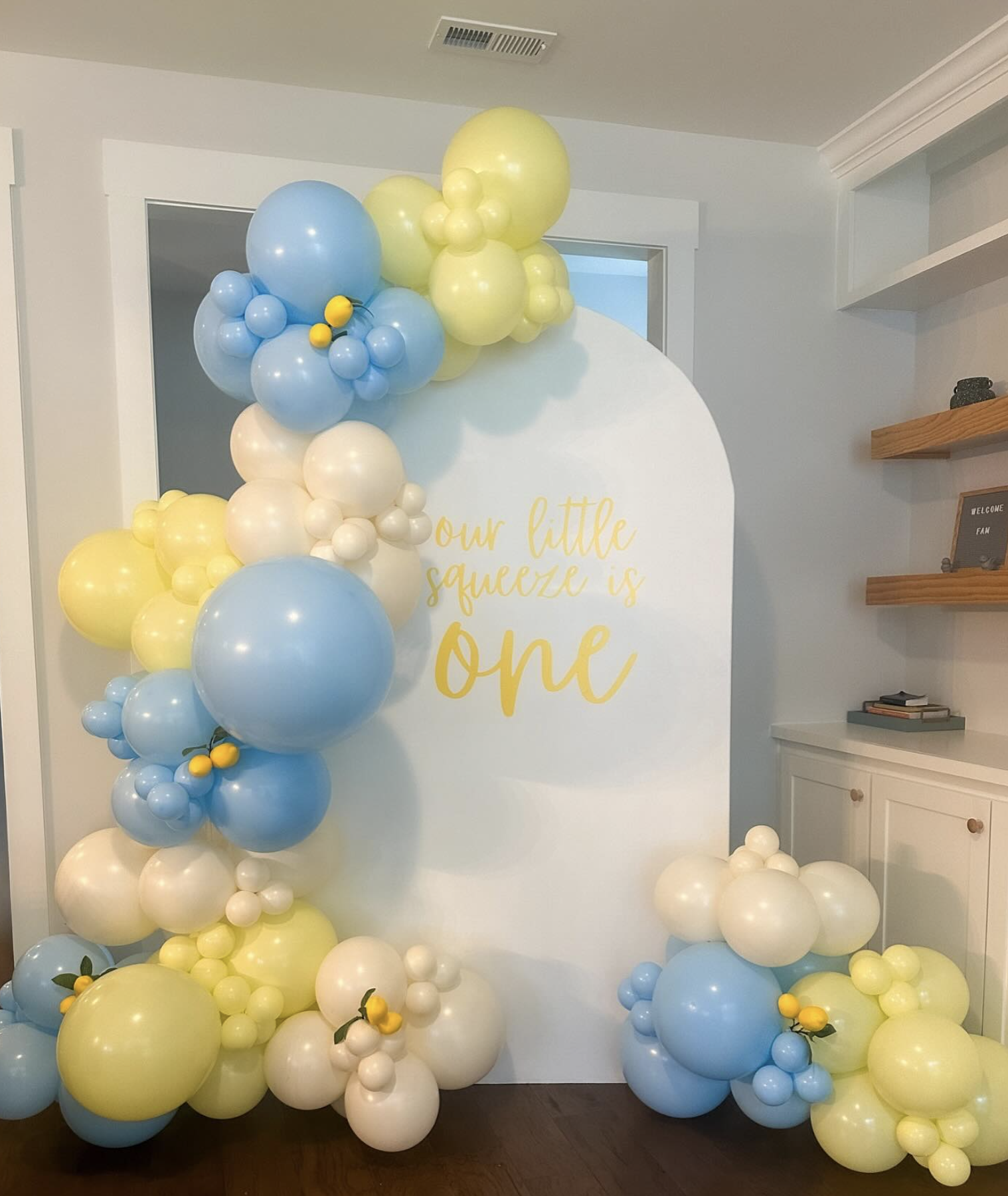 Decorative balloon arrangement with blue, yellow, and white balloons surrounding a white backdrop with the phrase 'our little squeeze is one' written in yellow.