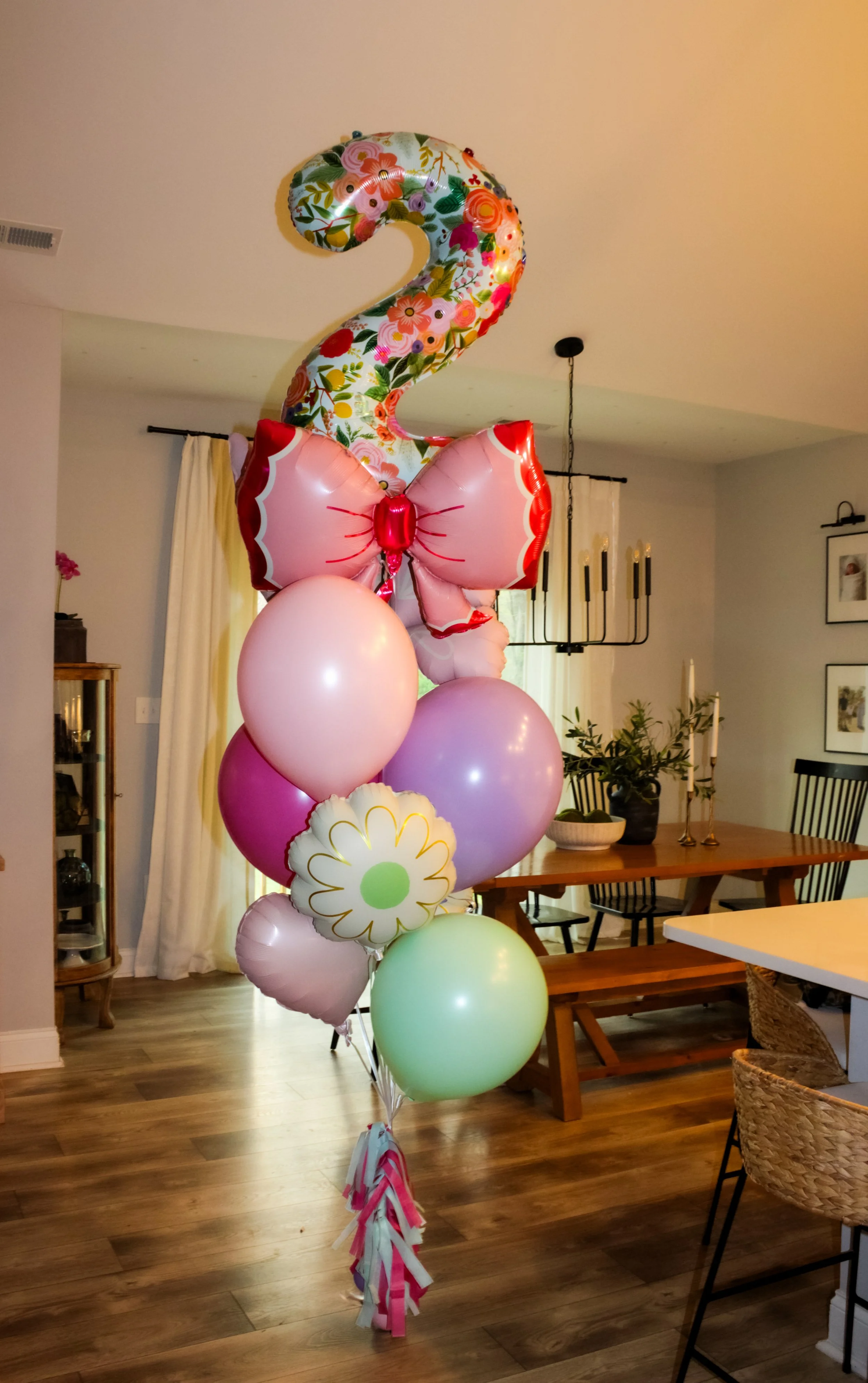 Colorful birthday decoration with a large number 2 foil balloon, pink and floral patterned, and a pink butterfly-shaped foil balloon, along with various pastel-colored balloons in a dining room.