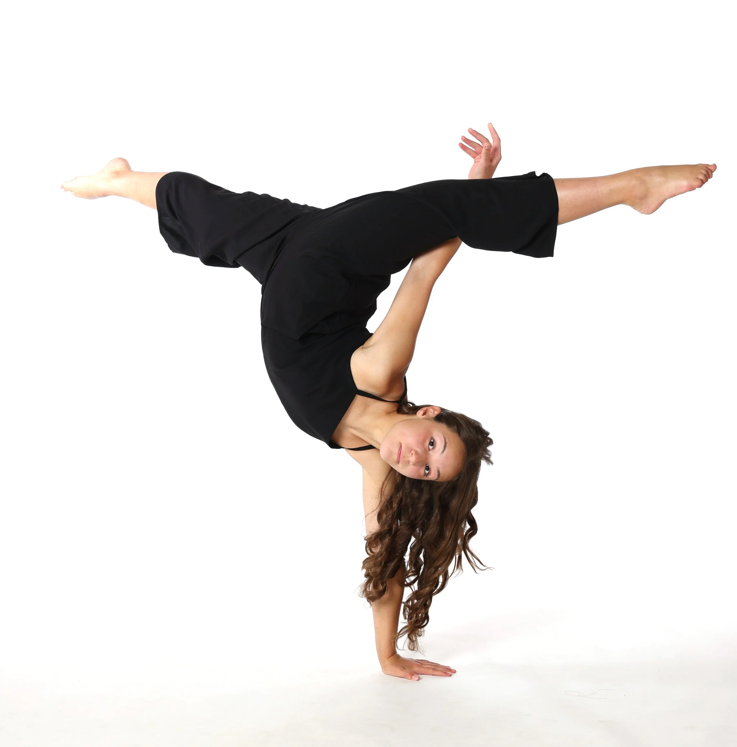 A woman performing a one-handed handstand, balancing on her right hand with her left arm extended outwards, wearing a black tank top and black pants, with long curly hair against a white background.