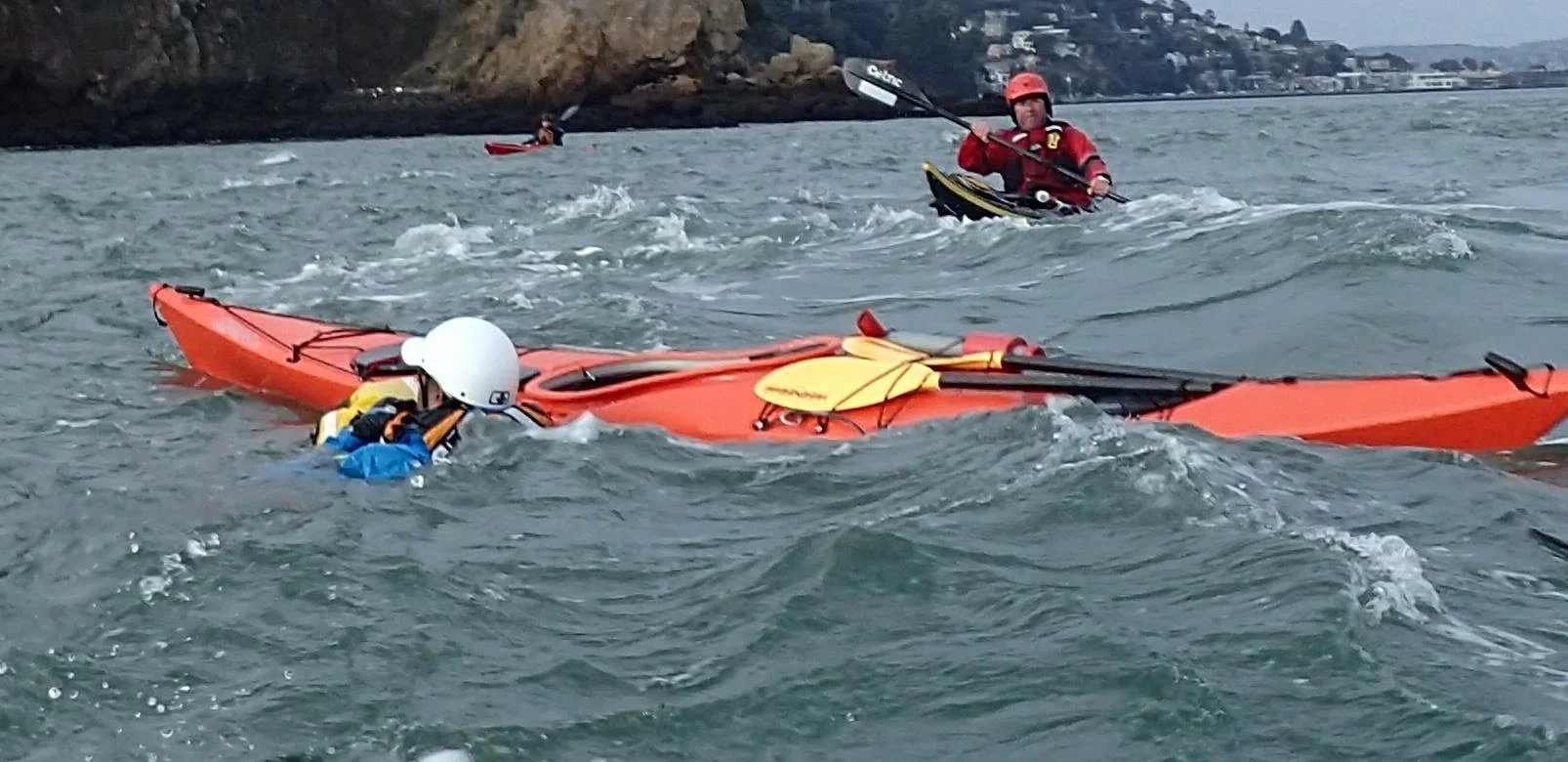 Kayakers in rough waters, one person in the water near an overturned kayak, others assisting.