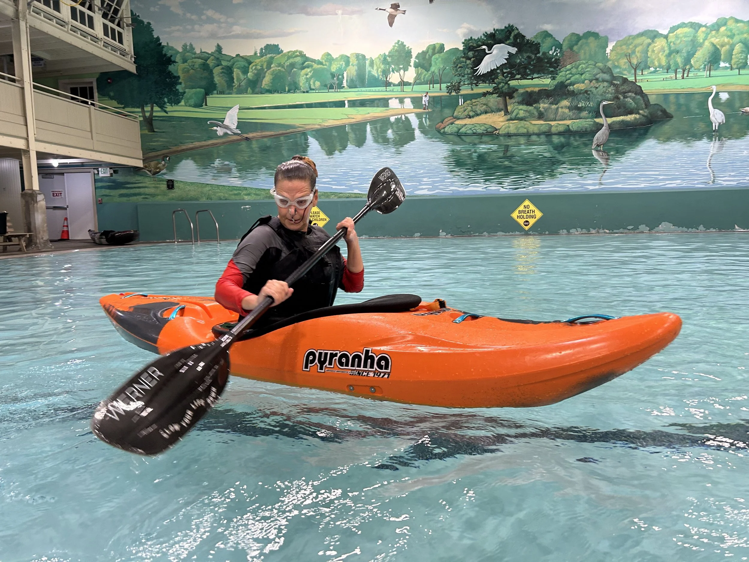 Person kayaking in indoor pool with mural background, holding a paddle and wearing goggles.