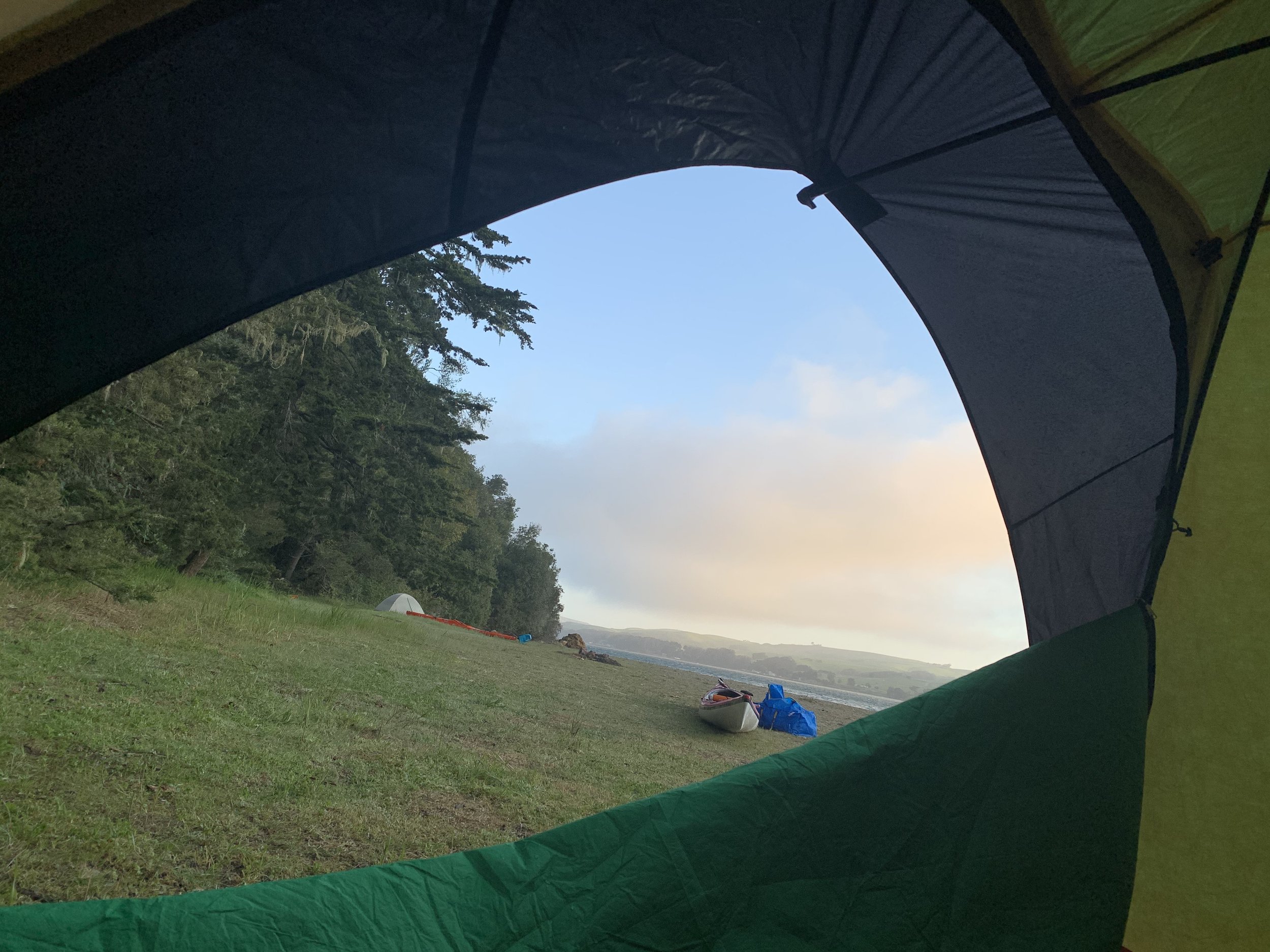View from inside a tent showing grassy field, trees, and a distant canoe.