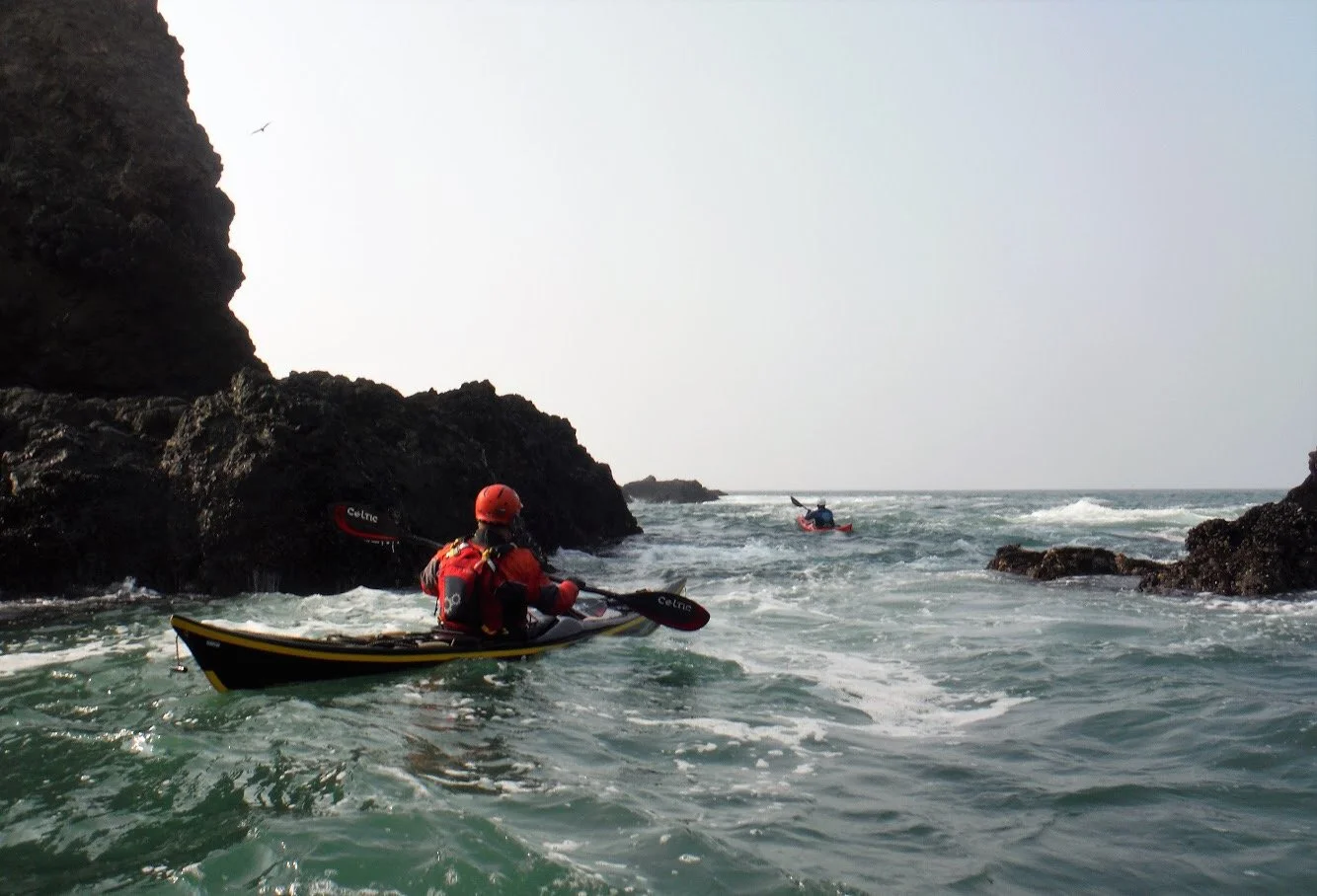 Two people kayaking near rocky coastline with waves