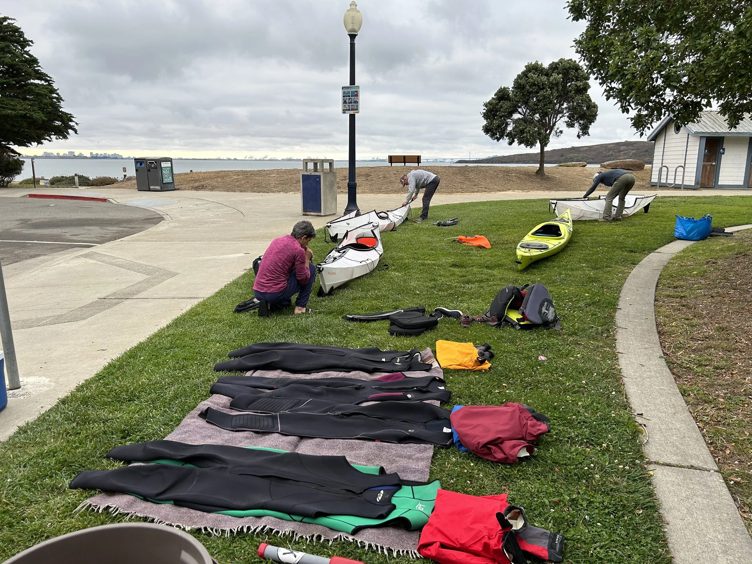 People preparing kayaks and wetsuits in a park area near a beach.
