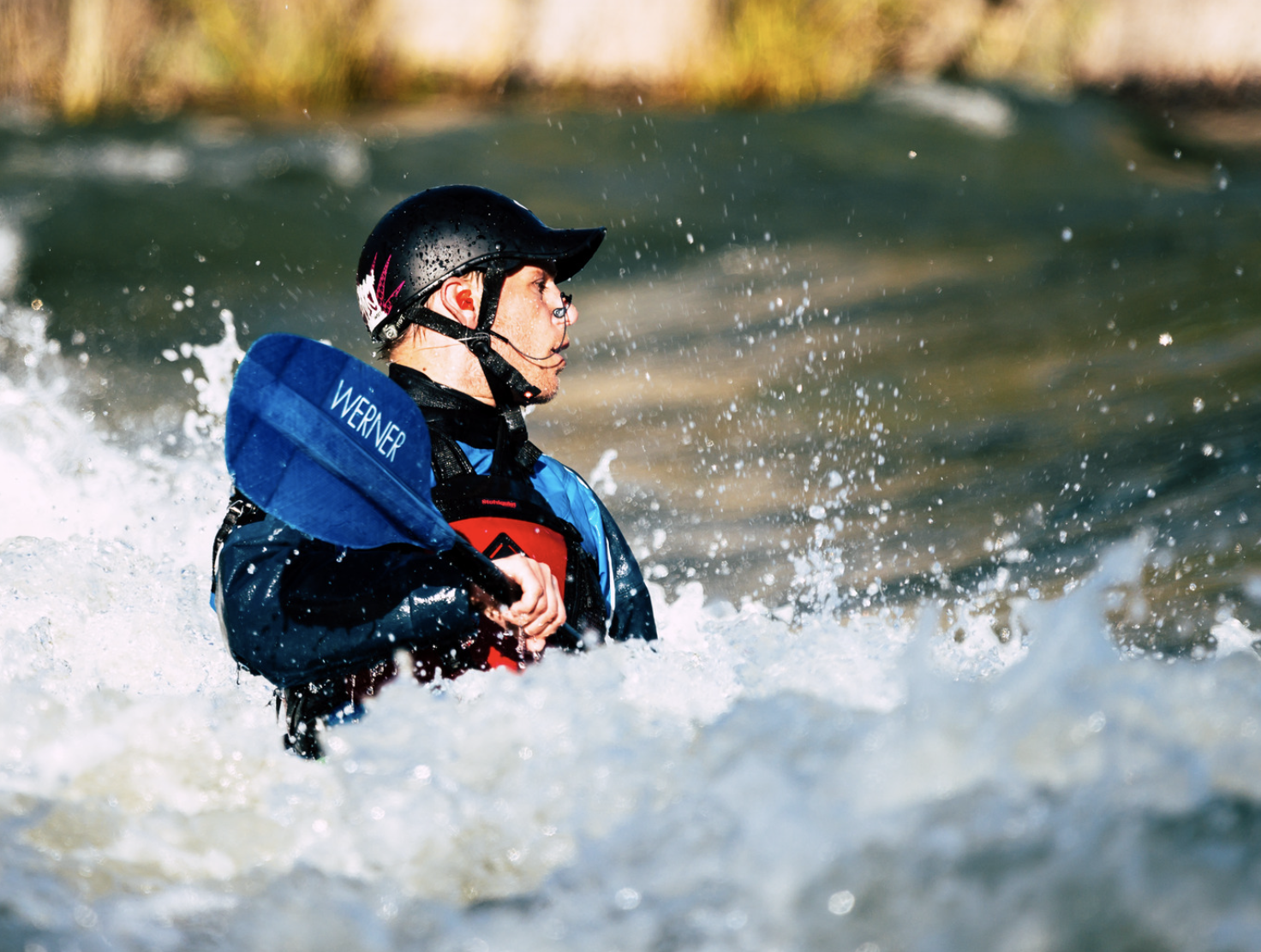 Person kayaking in rough river water wearing a helmet and life jacket holding a paddle.