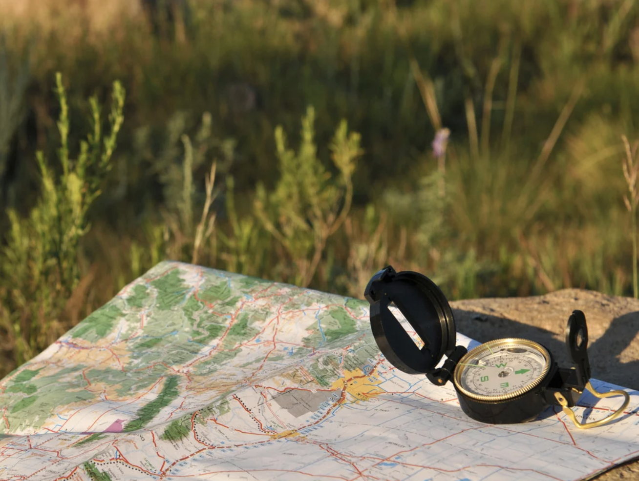 A compass placed on an unfolded map in an outdoor setting with green vegetation in the background.