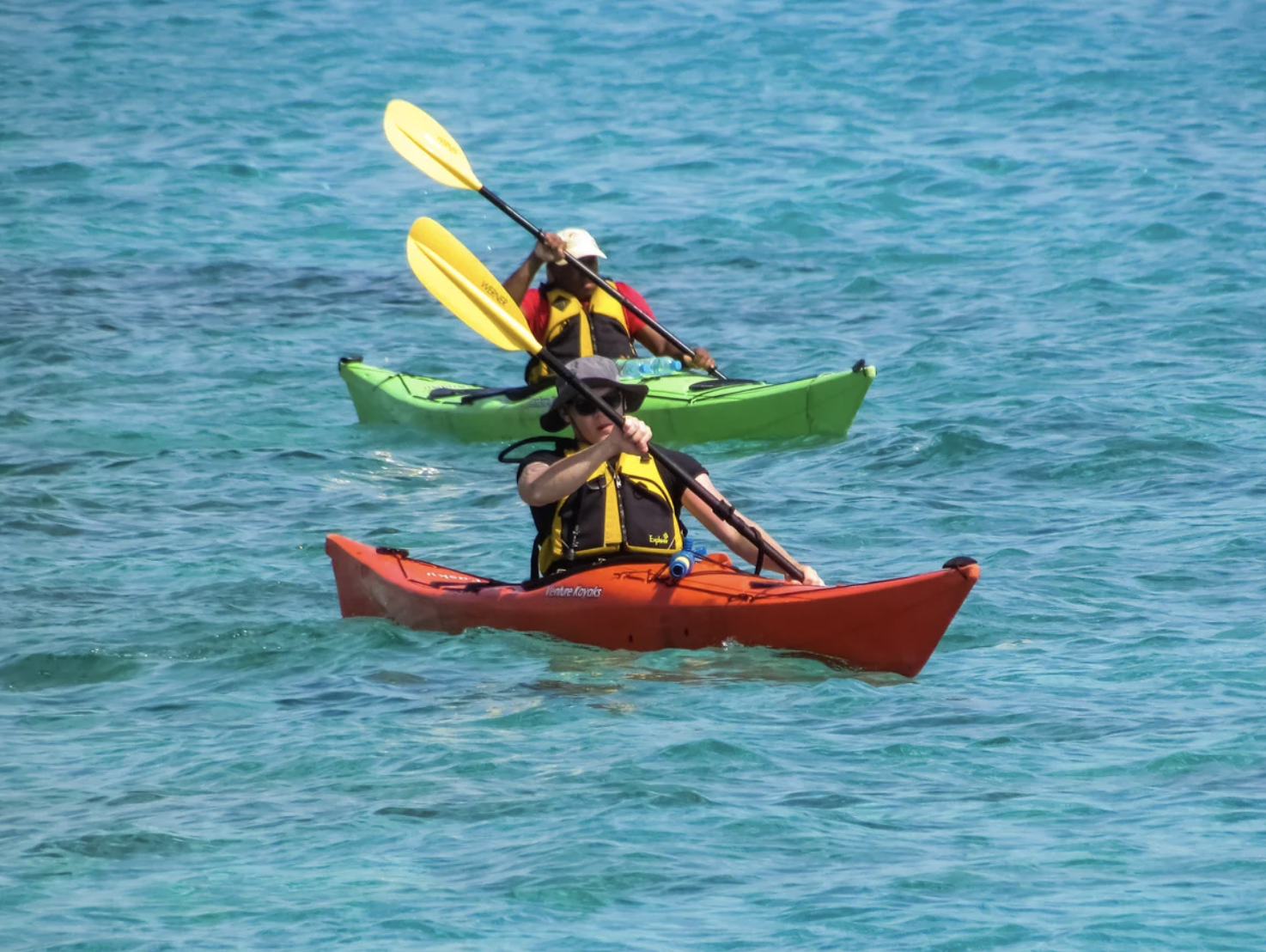 Two people kayaking on clear blue water, one in an orange kayak and the other in a green kayak, both wearing yellow life jackets.