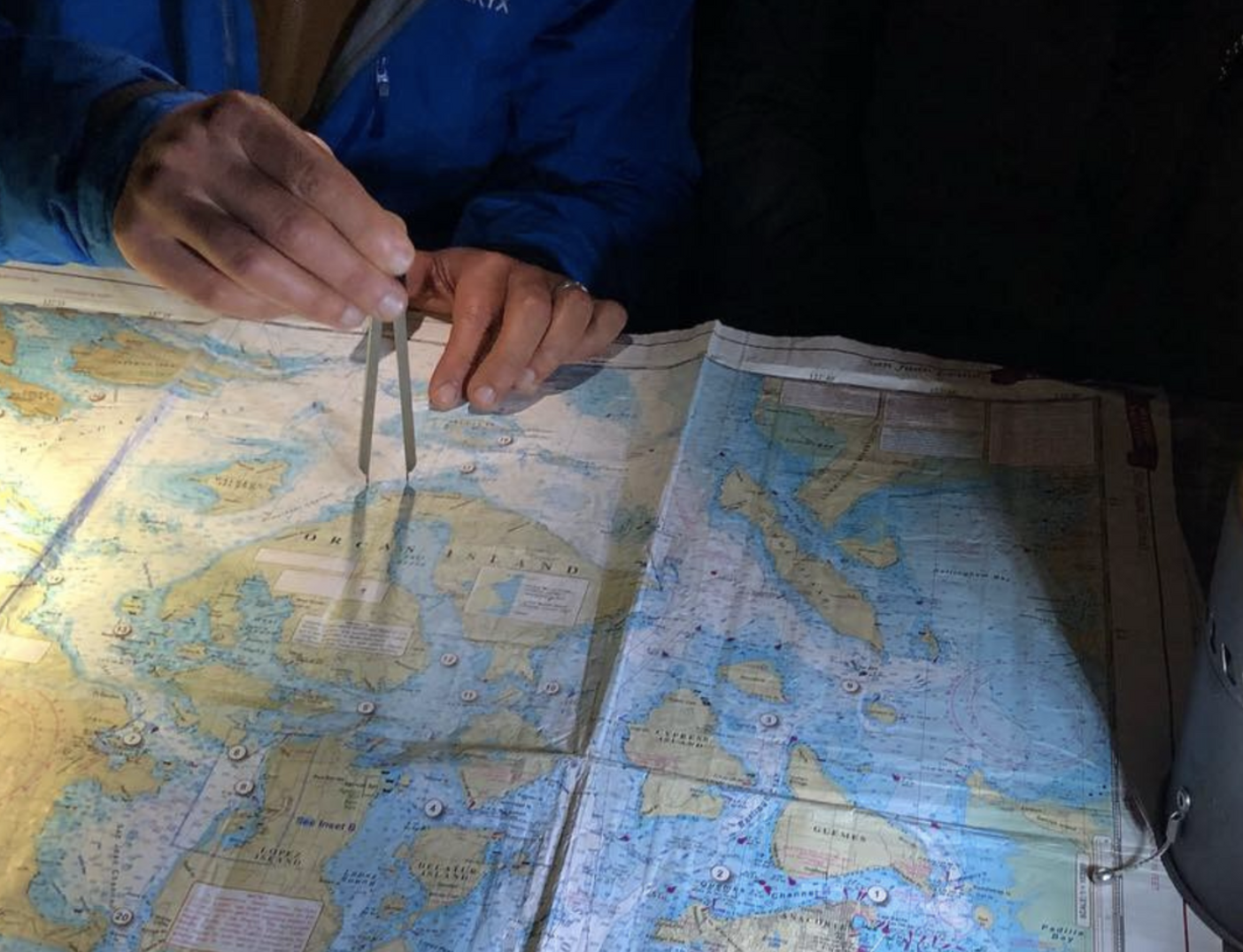 Person using a divider on a nautical map, wearing a blue jacket, dim lighting