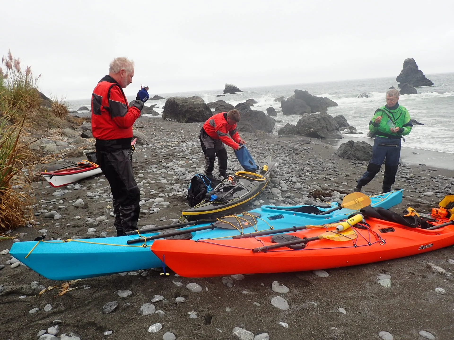 Three people on a rocky beach wearing kayaking gear, with kayaks in blue, orange, and yellow on the sand, near the ocean and large rocks.