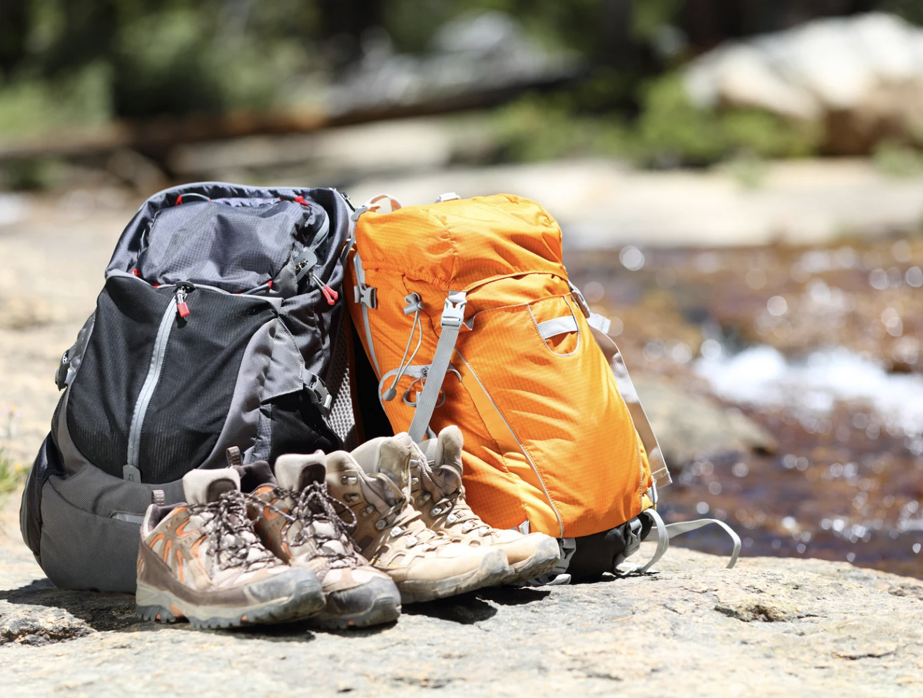 Two backpacks, one black and one orange, with two pairs of hiking boots on a rocky surface near a river.