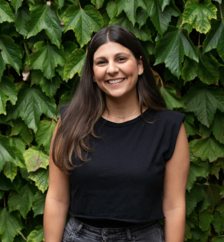 Woman smiling in front of green leafy background, wearing black sleeveless top and gray jeans.