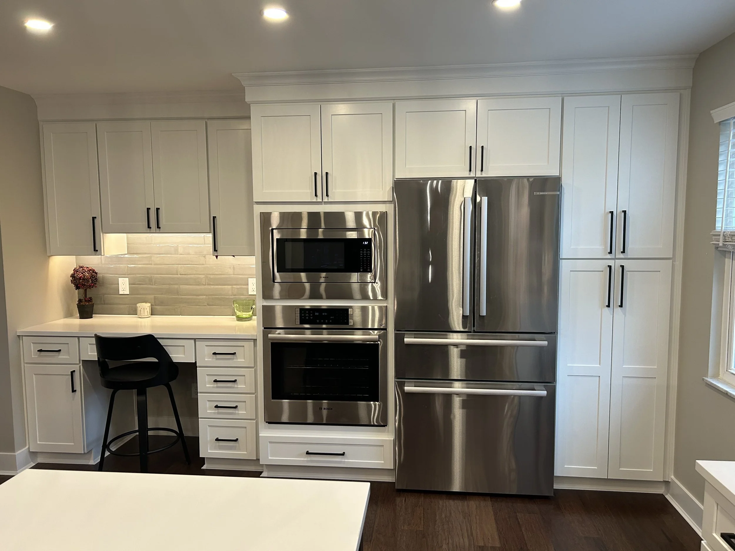 Modern kitchen with white cabinets, stainless steel appliances including a microwave, oven, and refrigerator, a small desk area with a black chair, and a window with blinds.