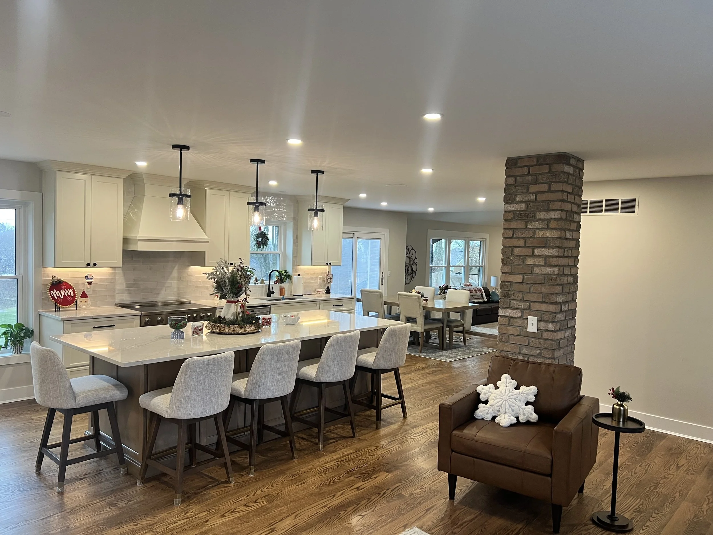 Open-concept living space with a kitchen, dining, and sitting area, decorated for Christmas. The kitchen has white cabinets, a large island with beige chairs, pendant lights, and holiday decor. The living area features a sitting chair with a snowflak