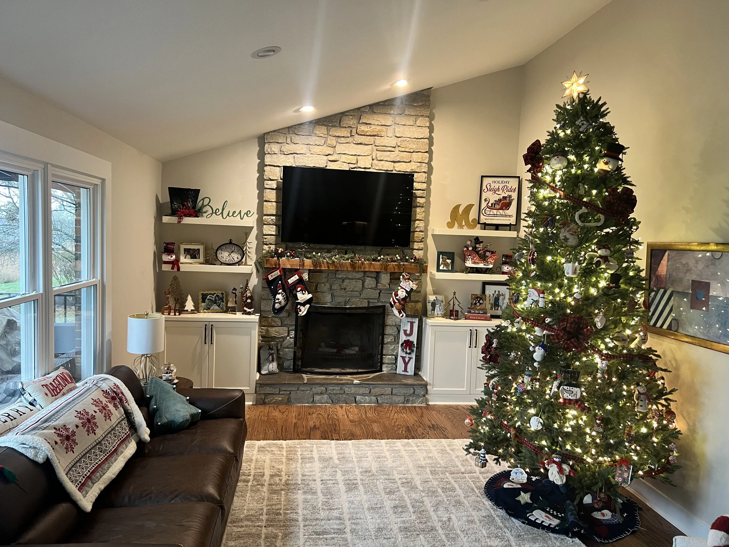 Living room decorated for Christmas with a lit Christmas tree on the right, a fireplace with stockings, and holiday decorations on shelves and furniture.