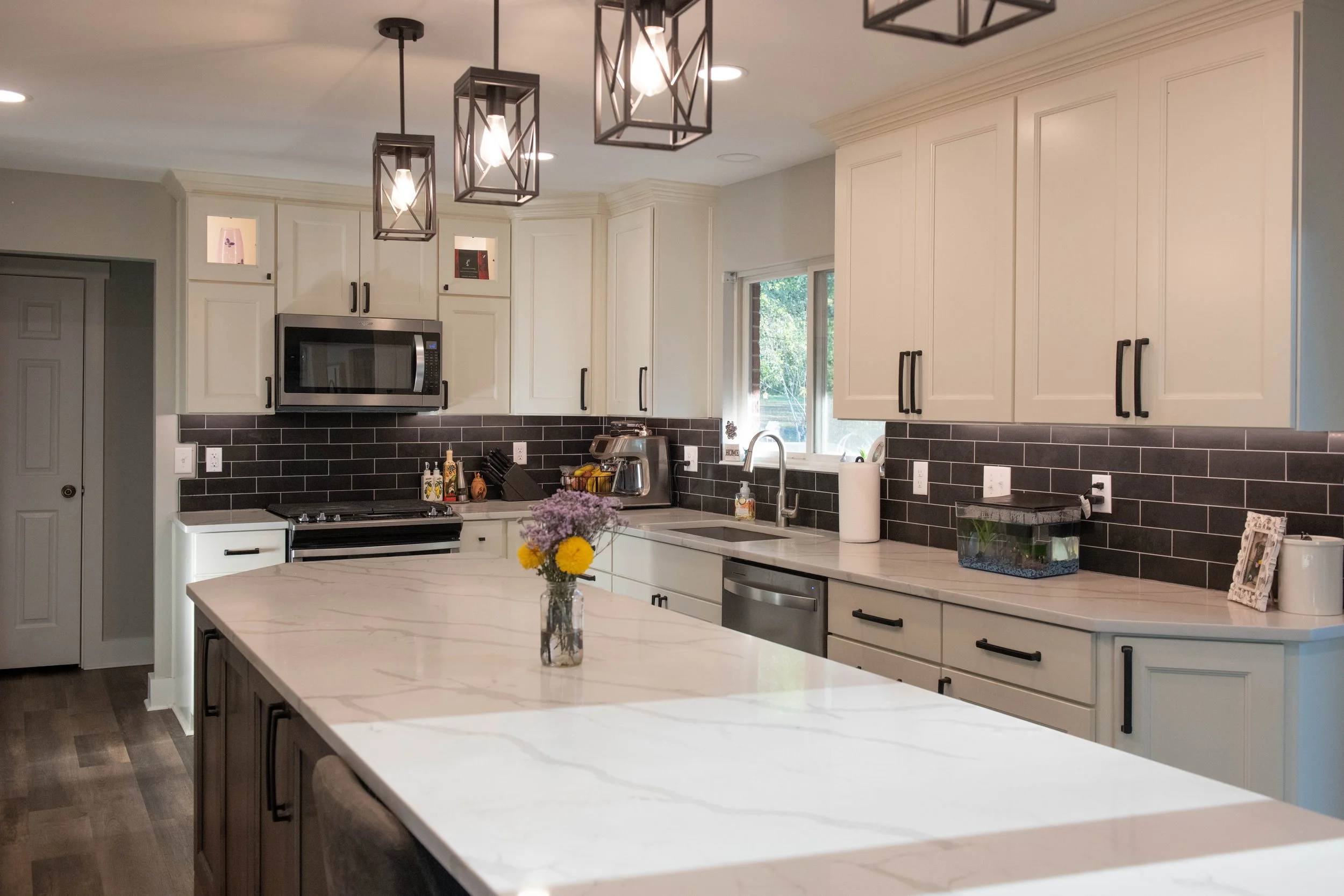Modern kitchen with white cabinetry, black hardware, black subway tile backsplash, marble countertop island, stainless steel appliances, and hanging geometric pendant lights.