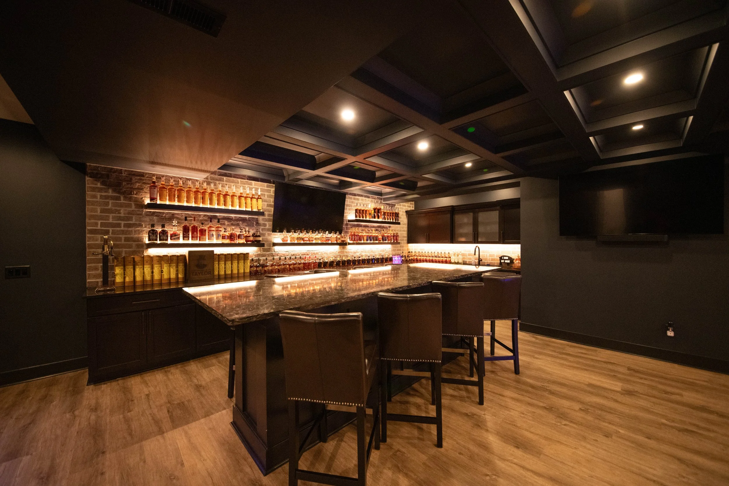 A dimly lit home bar with a dark granite countertop, wooden bar stools, exposed brick wall with backlit shelves holding bottles of spirits, a flat-screen TV, and a large TV on the wall, with hardwood flooring.