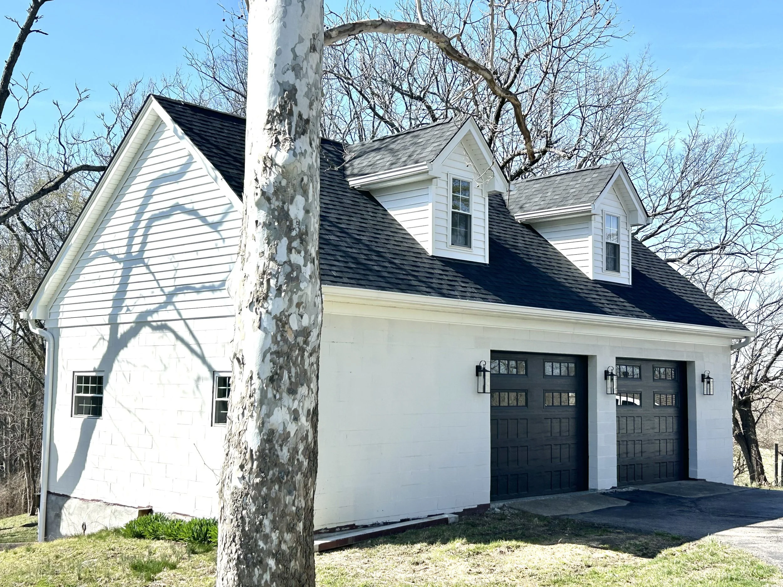 White house garage with two black garage doors and two dormer windows on the roof. A tree with a mottled bark is in the foreground, casting a shadow on the house. The sky is clear and blue.