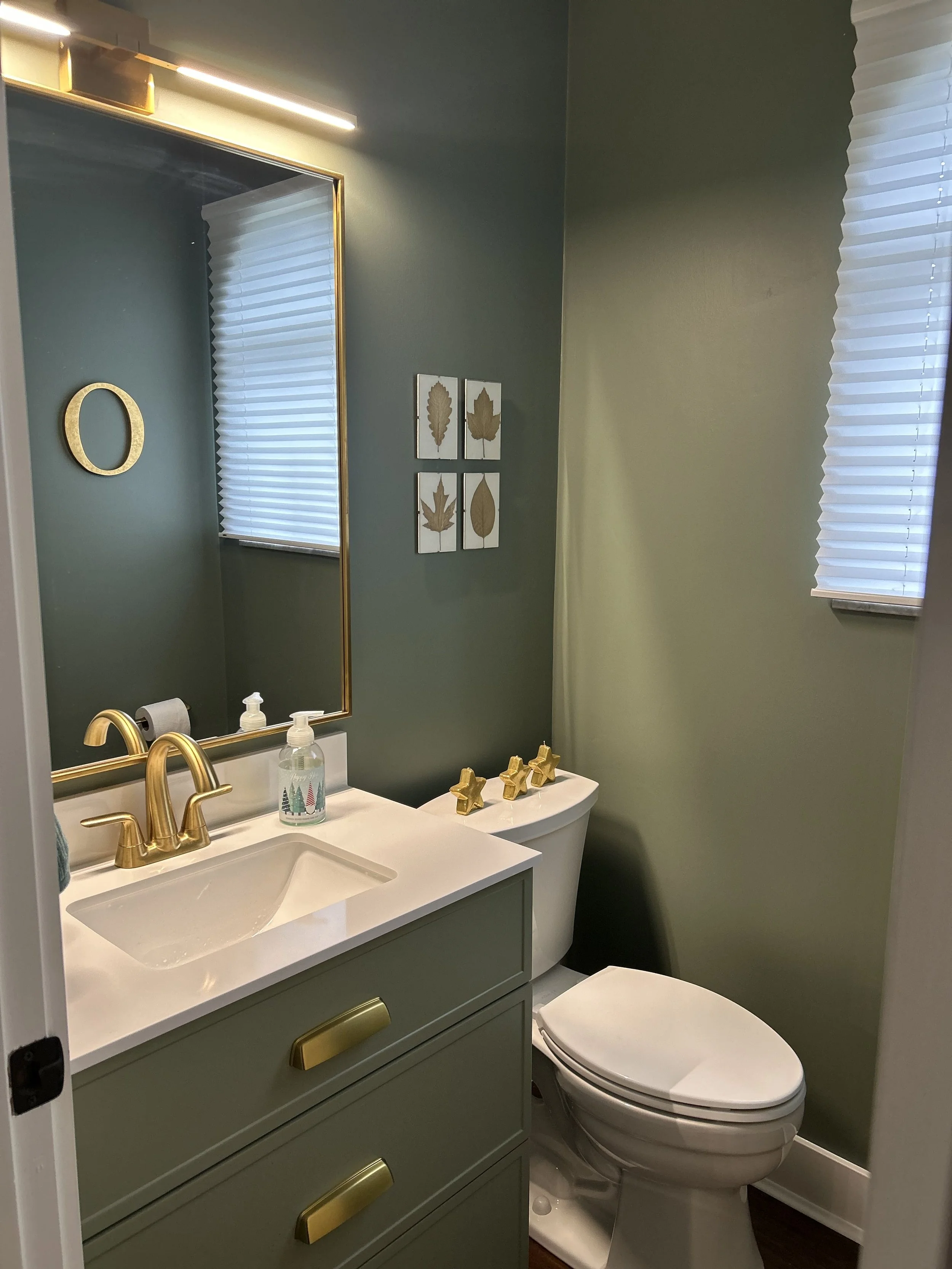 A small bathroom with green walls, a white vanity with gold handles, a porcelain sink, a mirror with a gold frame, and gold faucet fixtures. There are two small windows with white blinds, three framed leaf wall art pieces, and a white toilet with gol