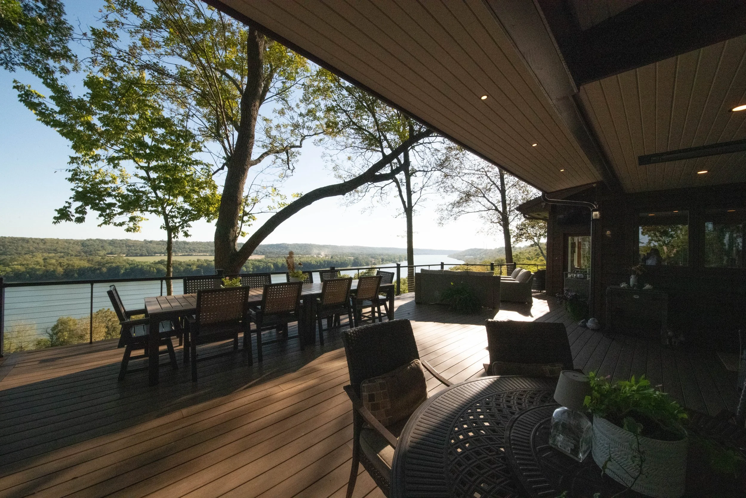 Outdoor deck with view of river and green trees, furnished with table, chairs, and lounge seating, under a covered roof with recessed lighting.