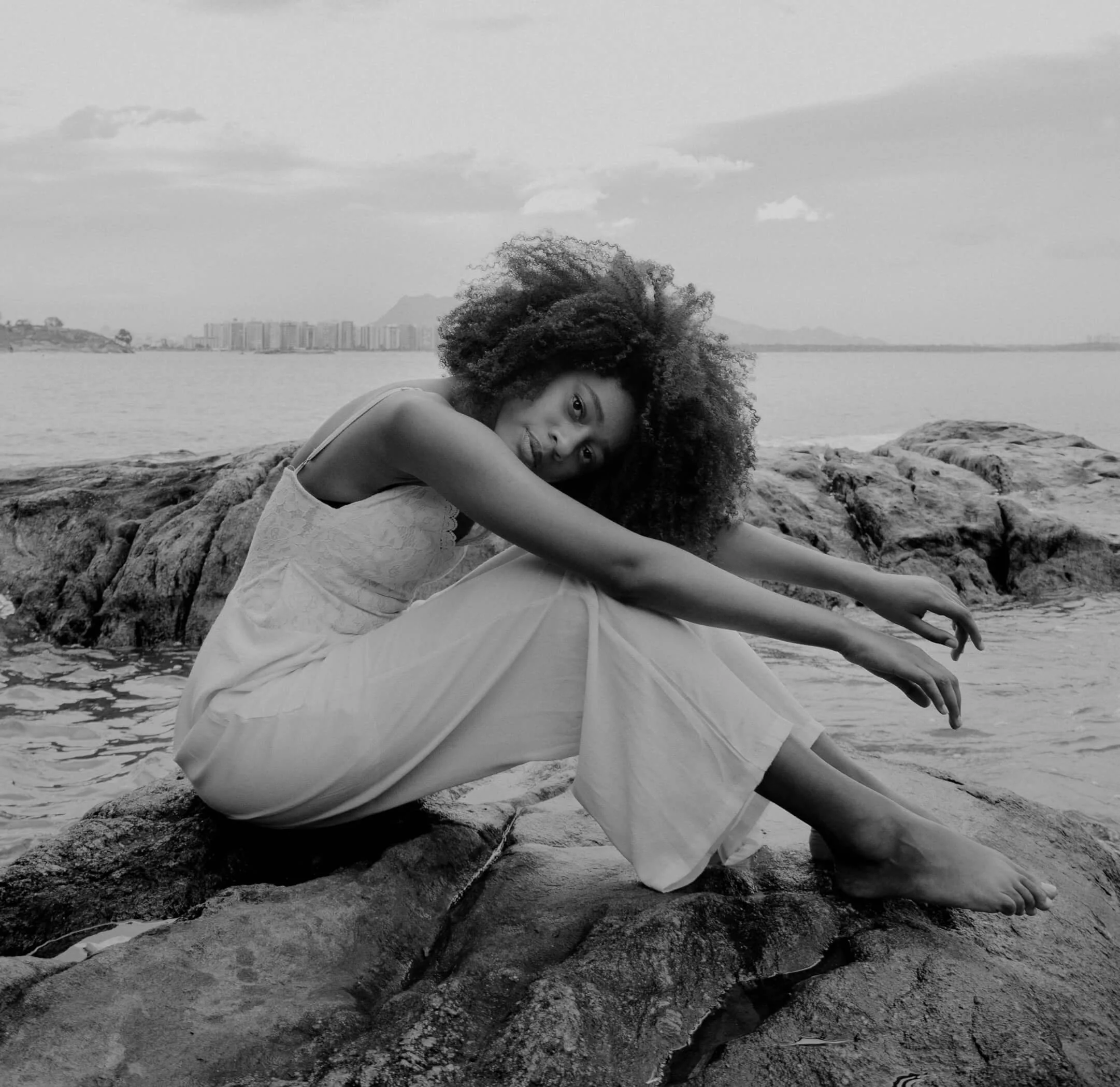 A woman sitting on rocks by the water with her arms extended, curly hair, wearing a light dress, with a cityscape and mountains in the background.
