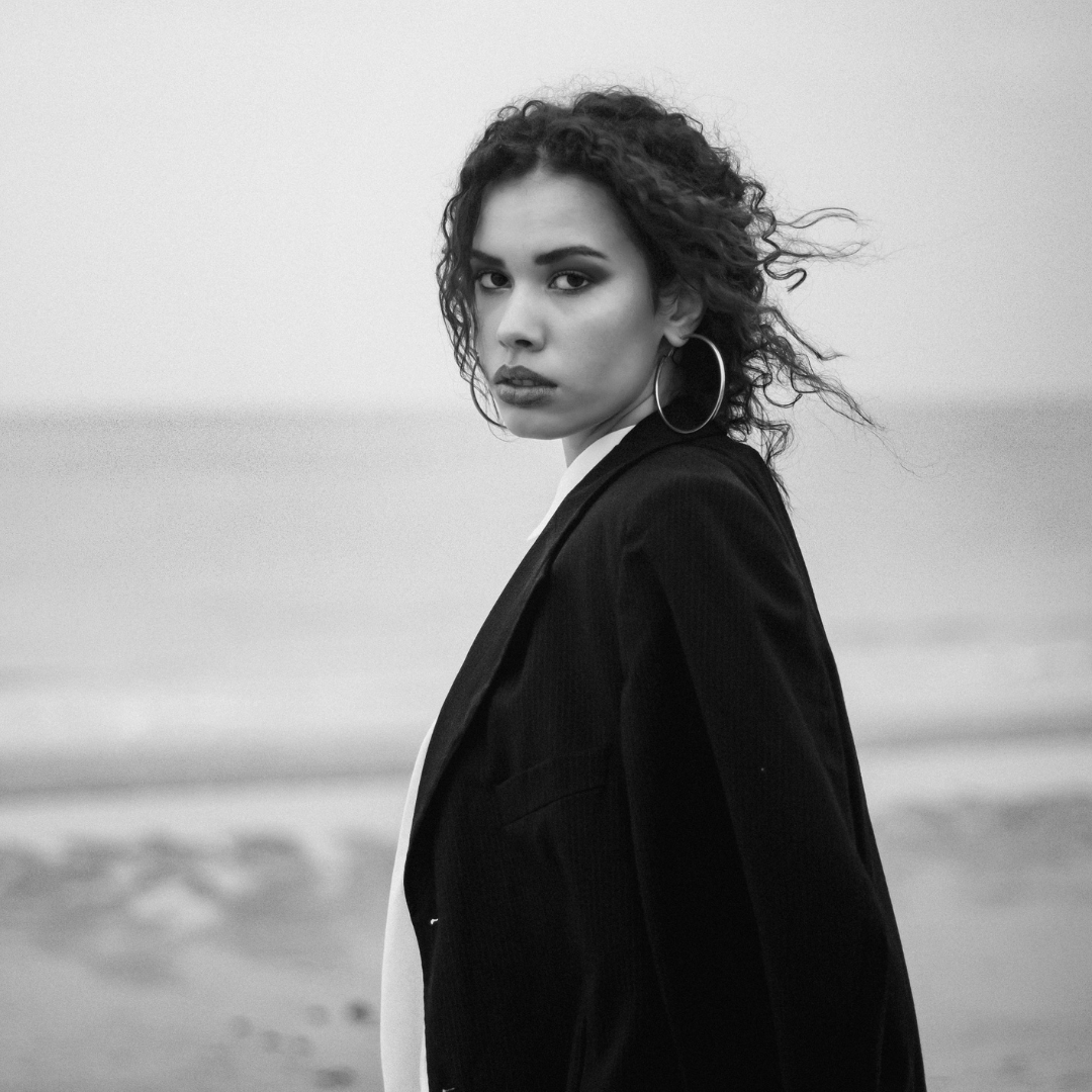 Black and white photo of a woman with curly hair wearing earrings and a dark blazer, standing by the beach.