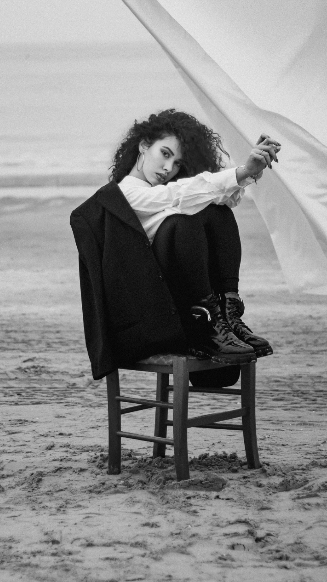 A woman sitting on a chair on the beach, holding a fabric or curtain, wearing a white shirt, black pants, and shiny black boots, with curly hair and hoop earrings.