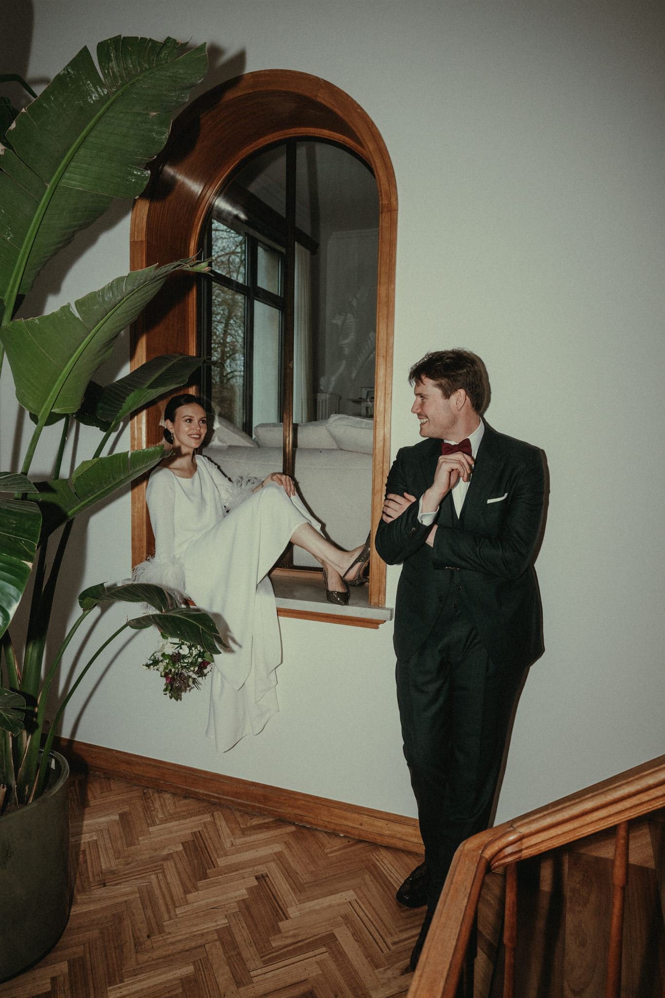 A woman in a white dress sitting on a ledge of a window, smiling at a man in a black tuxedo with a red bow tie, who is leaning against the wall and smiling back. Large green plants are in the foreground, and a wooden staircase is visible at the botto