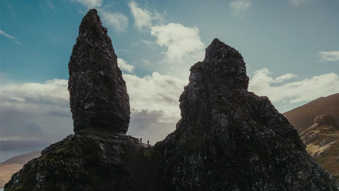Two large volcanic rock formations on a hillside with a cloudy sky in the background, and two small hikers walking near the base.