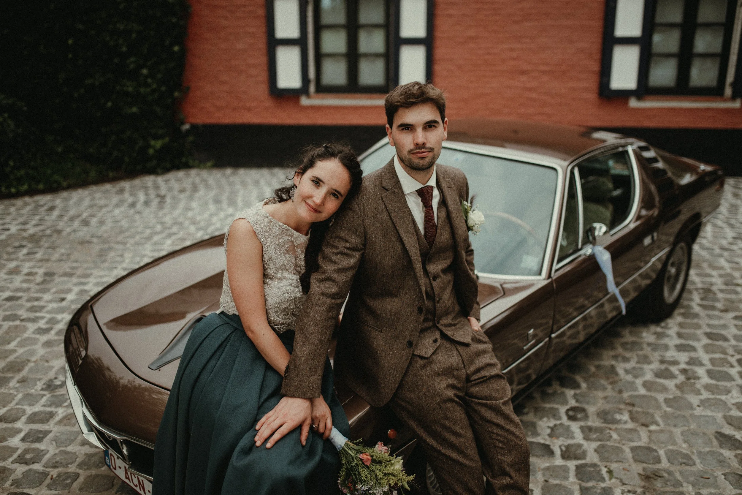 A bride and groom sitting on the hood of a vintage car in front of a brick house with windows. The bride is wearing a lace top and a teal skirt, leaning her head on the groom's shoulder. The groom is dressed in a brown suit with a tie. The bride is holding a bouquet of flowers.