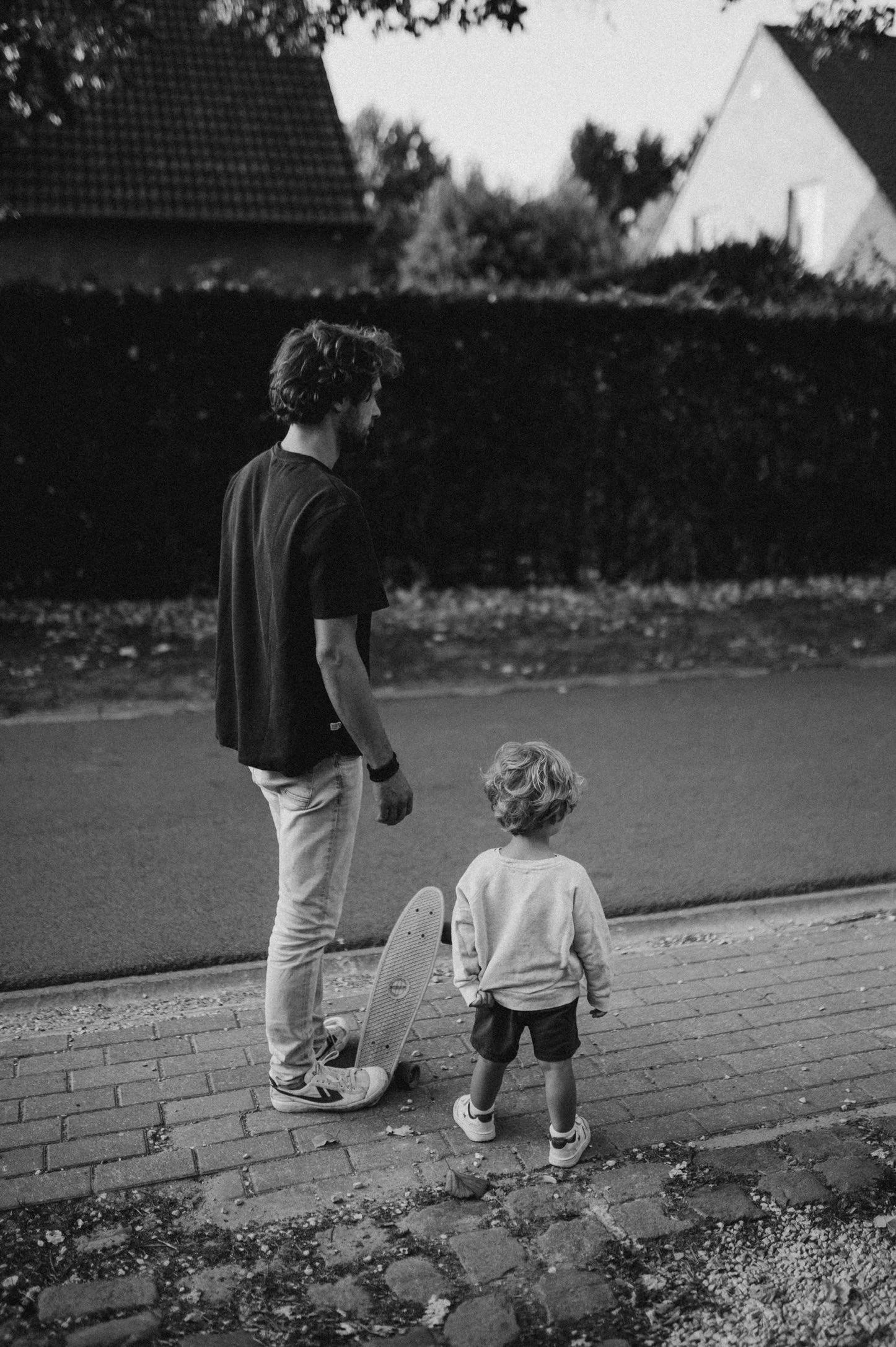 A black and white photo of an adult and a child standing on a sidewalk, with the child holding a skateboard. The adult has curly hair and is wearing a dark t-shirt and light pants. The child has short hair, a light sweatshirt, and shorts. They are facing away from the camera, looking at the street, with houses and trees in the background.