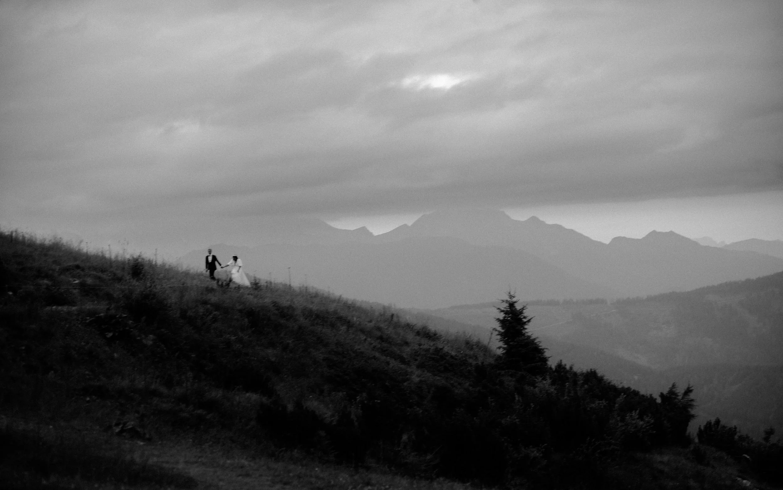 Black and white photo of a couple in wedding attire walking hand in hand on a hillside with mountains in the background and a cloudy sky overhead.