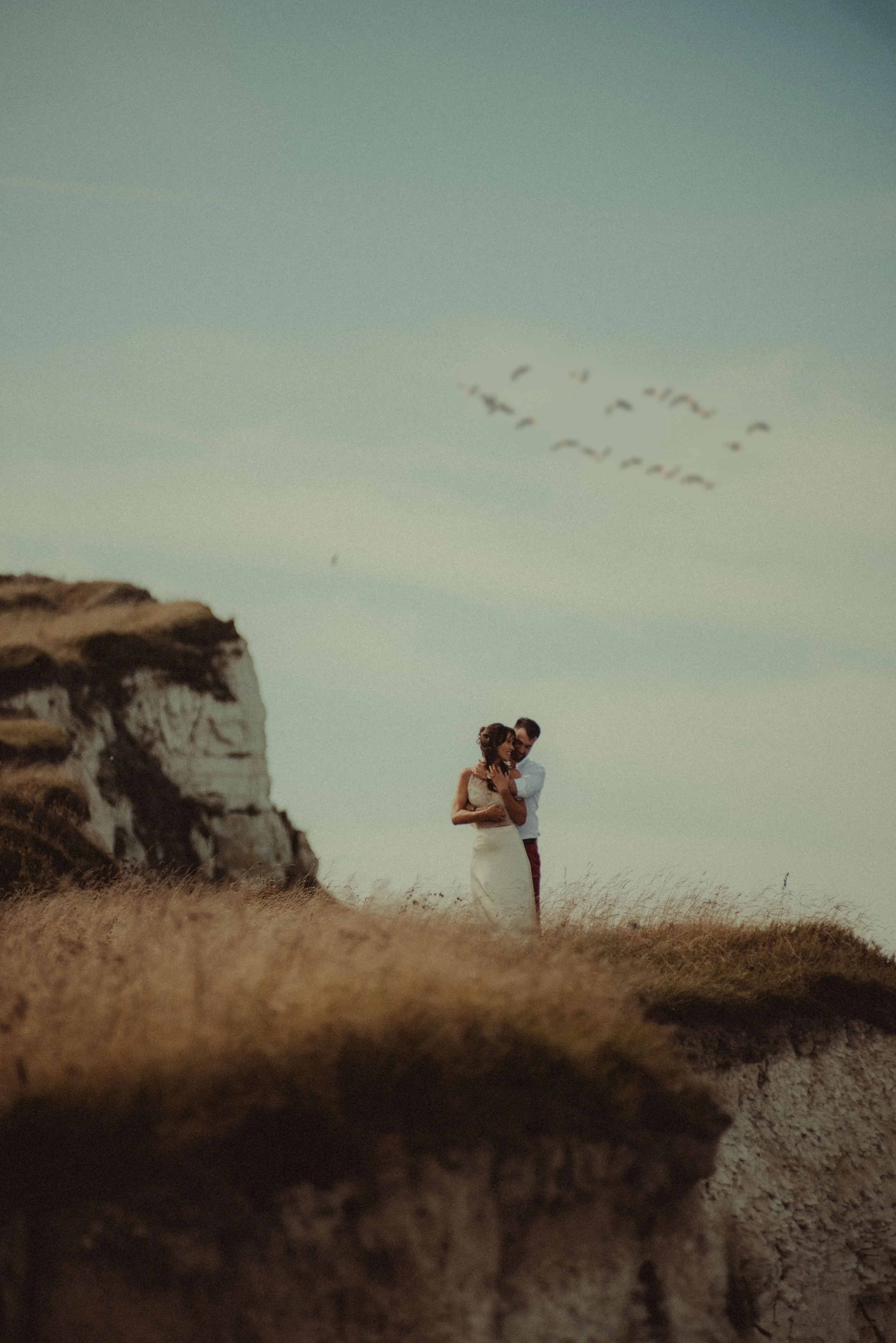 A couple standing on a grassy cliff during sunset, with a flock of birds flying overhead in a V formation.